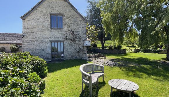 Stone cottage with large windows, surrounded by lush greenery. A wooden bench and table sit on the lawn under a clear blue sky.