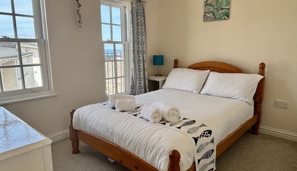 Cozy bedroom with a wooden bed, white linens, and rolled towels. Two windows offer a view of the sea. A blue lamp and wall art add color.