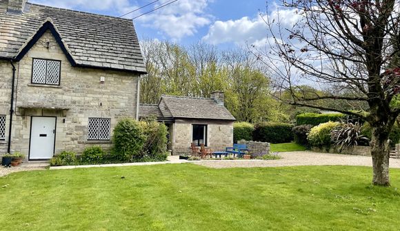 Stone cottage with a slate roof, surrounded by a lush green lawn, trees, and a gravel path under a partly cloudy sky.