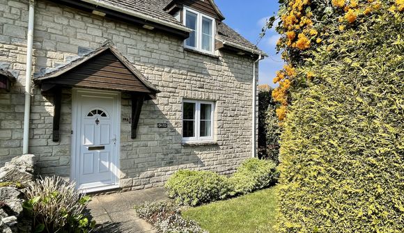 Stone cottage with a white door, small garden, and flowering hedge under a clear blue sky.