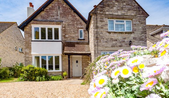 Stone house with large windows, surrounded by a gravel path and colorful daisies in the foreground, under a clear blue sky.