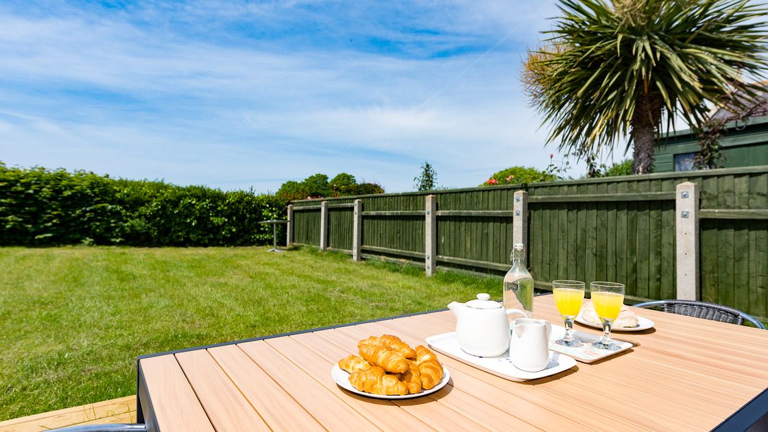 Outdoor table with croissants, orange juice, and tea set on a sunny day. Green lawn and palm tree in the background.