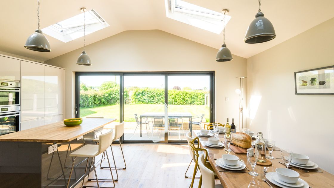 Bright kitchen with skylights, a wooden dining table set for six, island with stools, and large glass doors opening to a garden view.