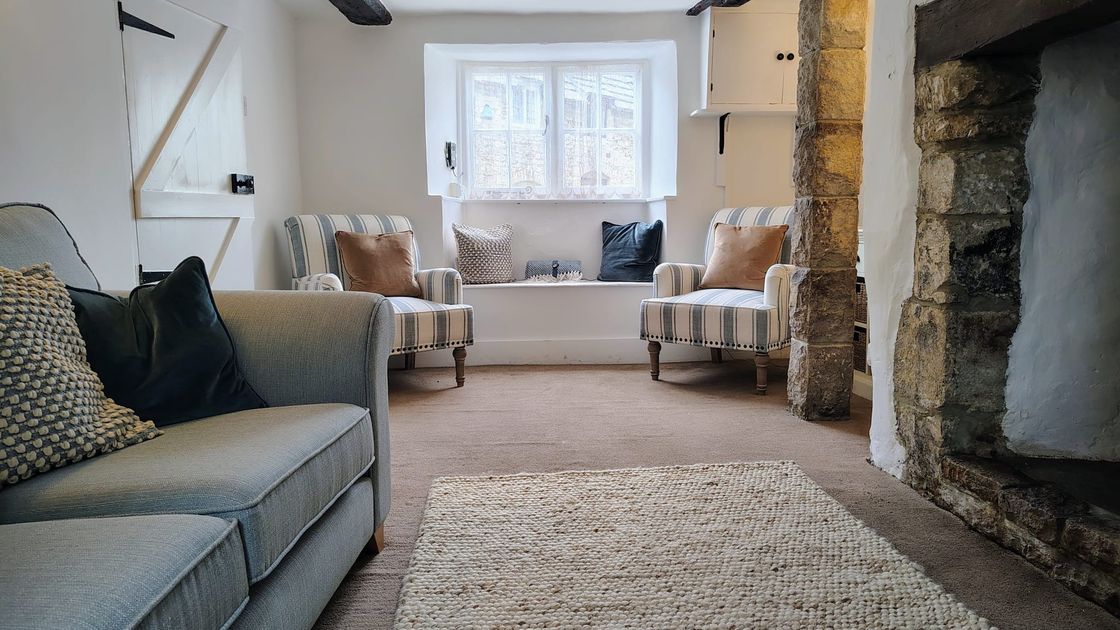 Cosy living room with a beige sofa, striped armchairs, stone fireplace, and a window seat. Neutral tones and rustic beams add charm.