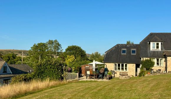 Stone house with a sloped roof, patio furniture, and a garden. Sunlit lawn and umbrella-covered table in the foreground. Clear blue sky.