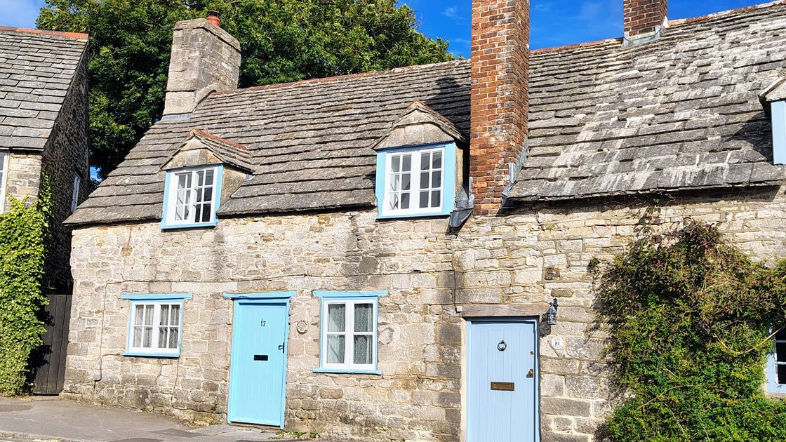 A quaint stone cottage with a blue door and window frames, featuring a sloped roof and chimney against a clear blue sky.