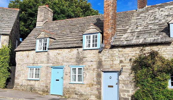 A quaint stone cottage with a blue door and window frames, featuring a sloped roof and chimney against a clear blue sky.