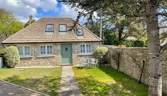 Charming stone cottage with a green door, white-framed windows, and a small patio set in a grassy yard, surrounded by trees and a stone wall.
