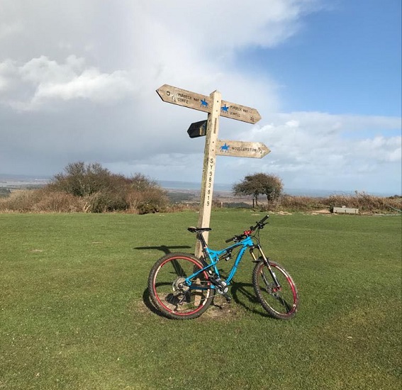 A mountain bike leans against a wooden signpost on a grassy hilltop, with a vast landscape and cloudy sky in the background.