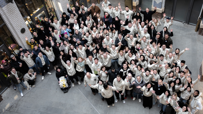 “Large group of smiling Eye-Able team members standing in a courtyard, waving up at the camera, many wearing matching light-colored hoodies
