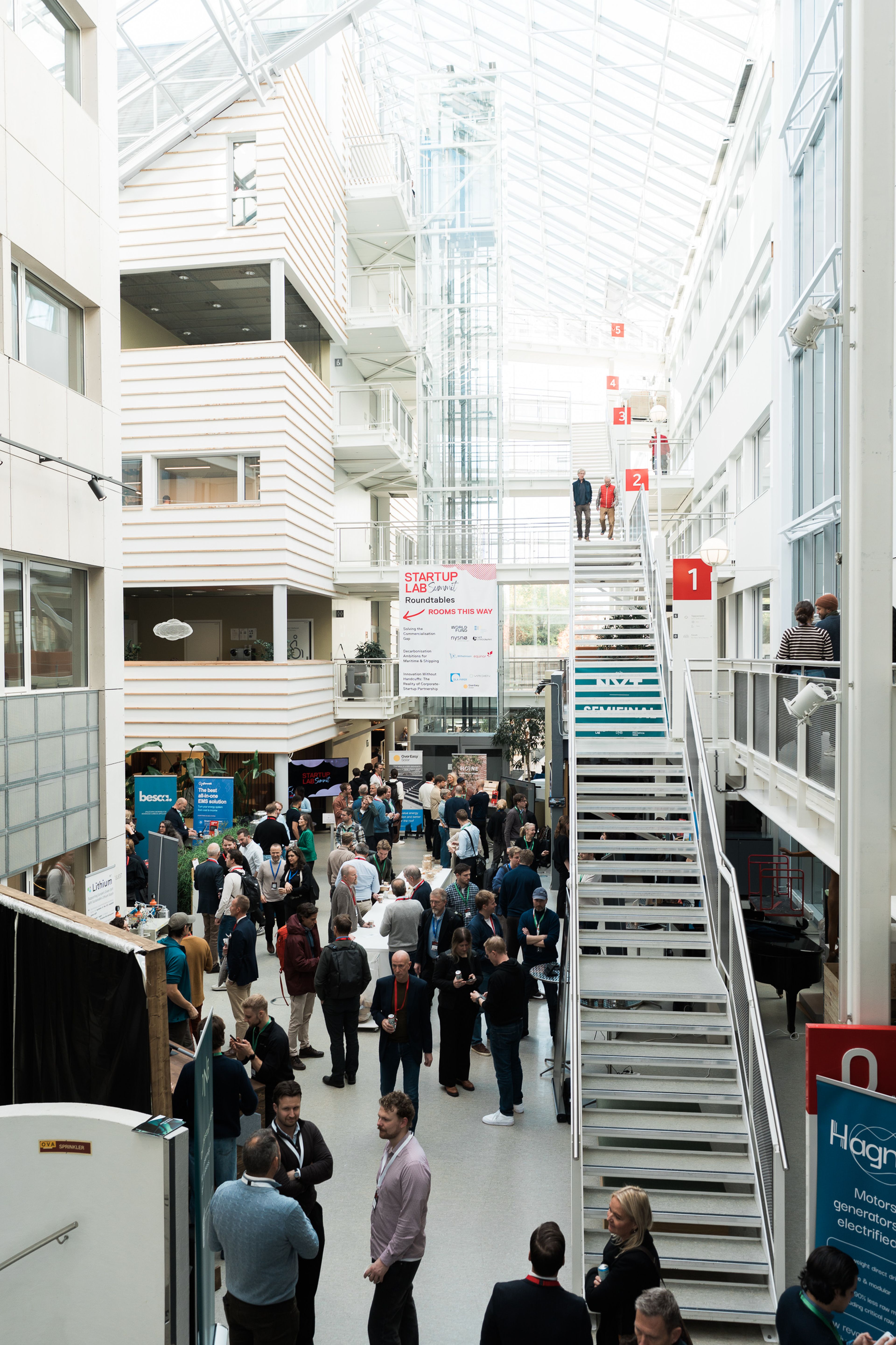 Interior of a modern building with a large crowd of people networking and attending an event, surrounded by banners and exhibits.