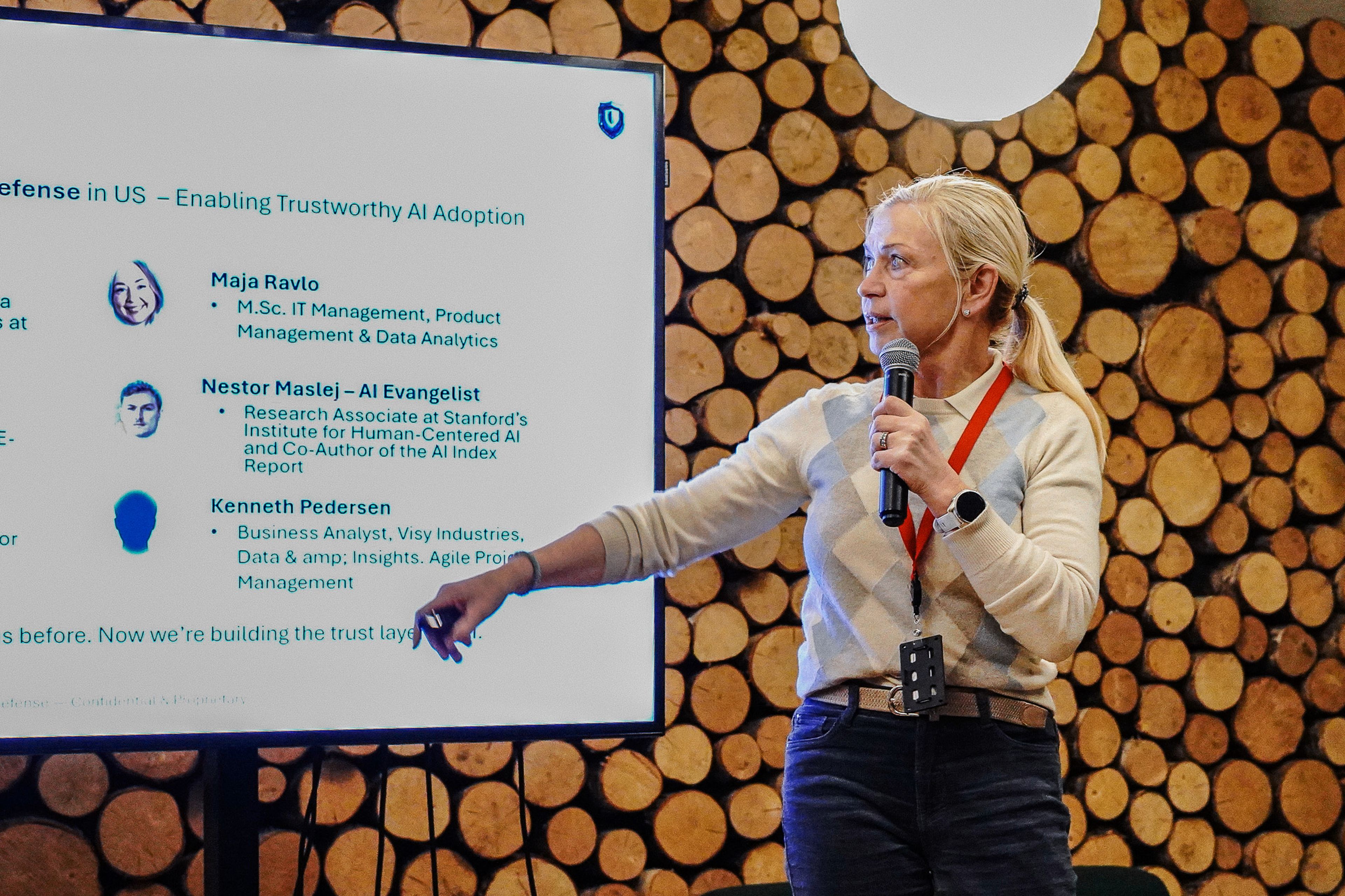 A woman with a microphone points to a presentation slide on a screen. The background features stacked logs.