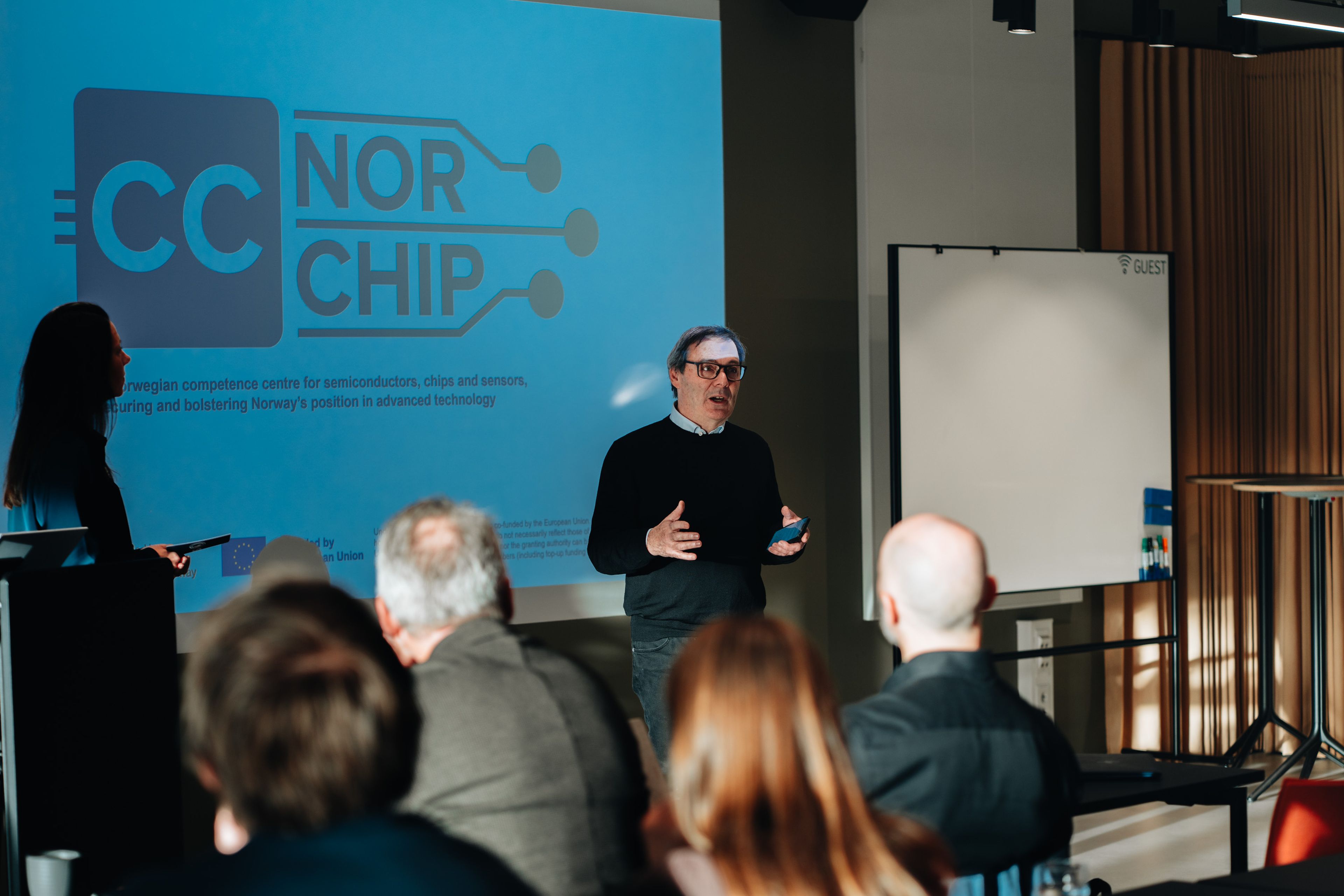 A person speaking in front of a projected presentation slide labeled "Norchip" with an audience seated in a conference room.