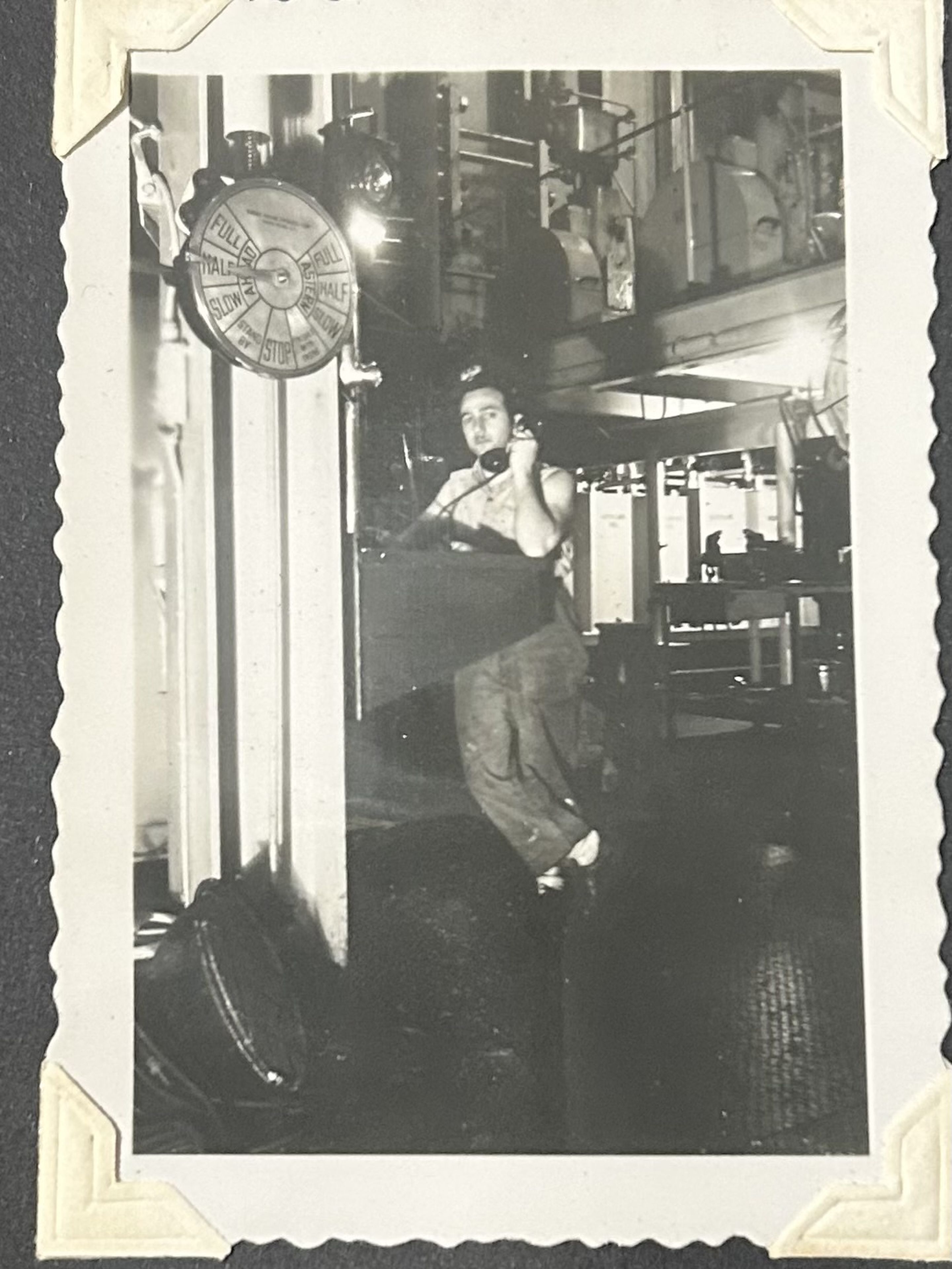 Joe in work attire stands on a ship, holding a phone. A telegraph and machinery are visible in the background. Black and white photo.