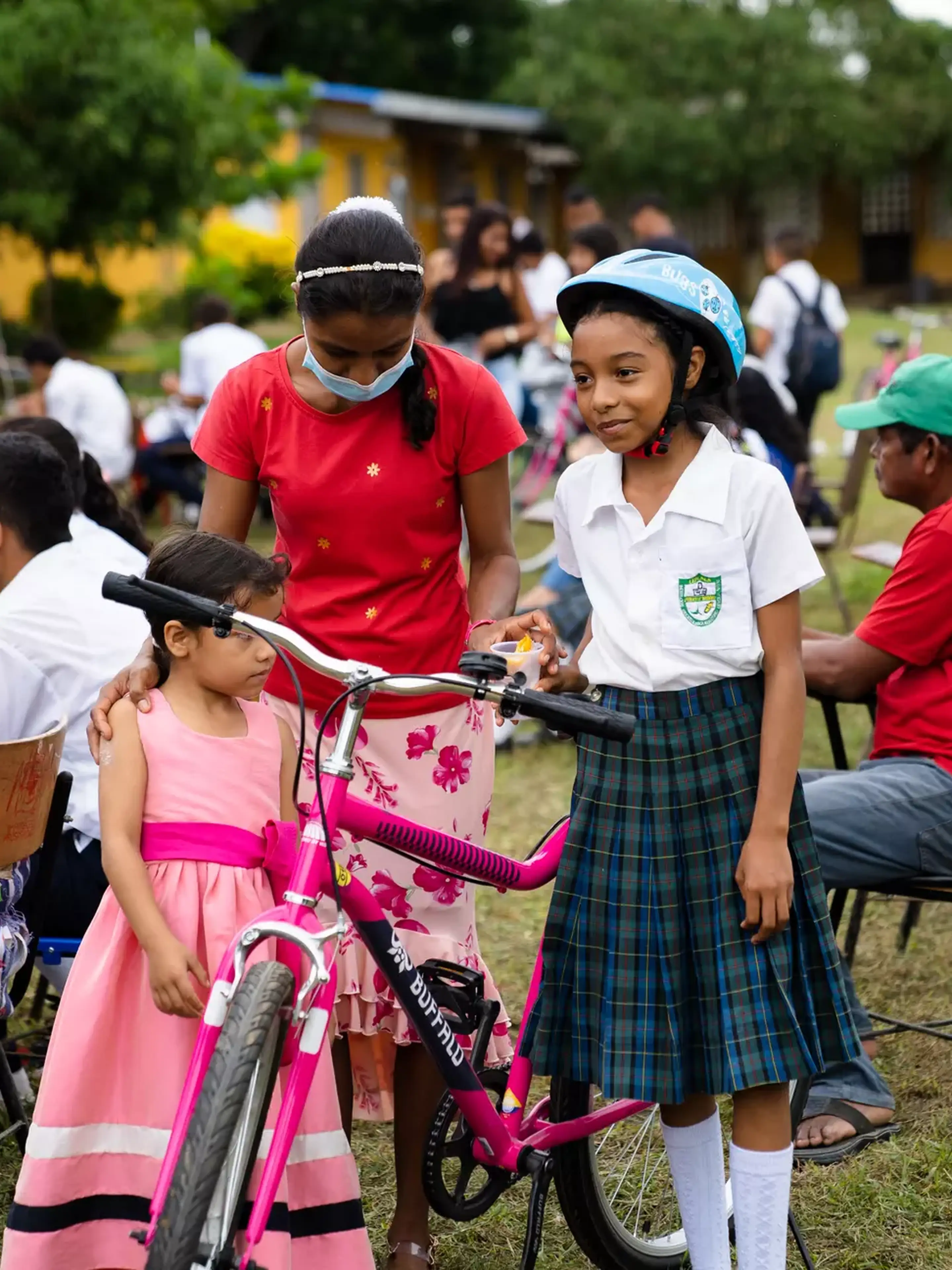 Girl in school uniform with bike helmet stands beside a pink bicycle, accompanied by a younger girl in a dress and a woman in a red top.