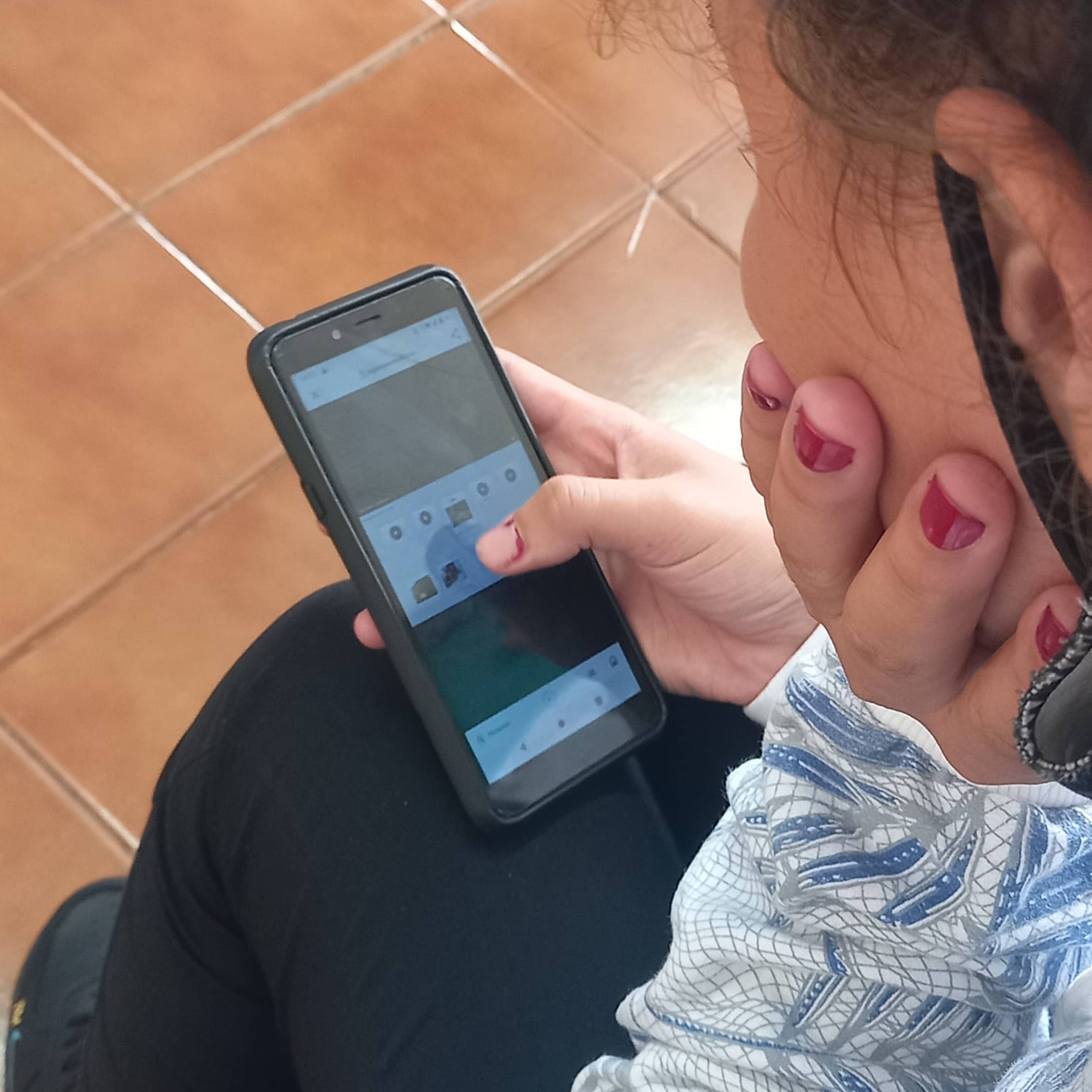 Person with pink nails using a smartphone, sitting on a tiled floor, browsing a language learning app.