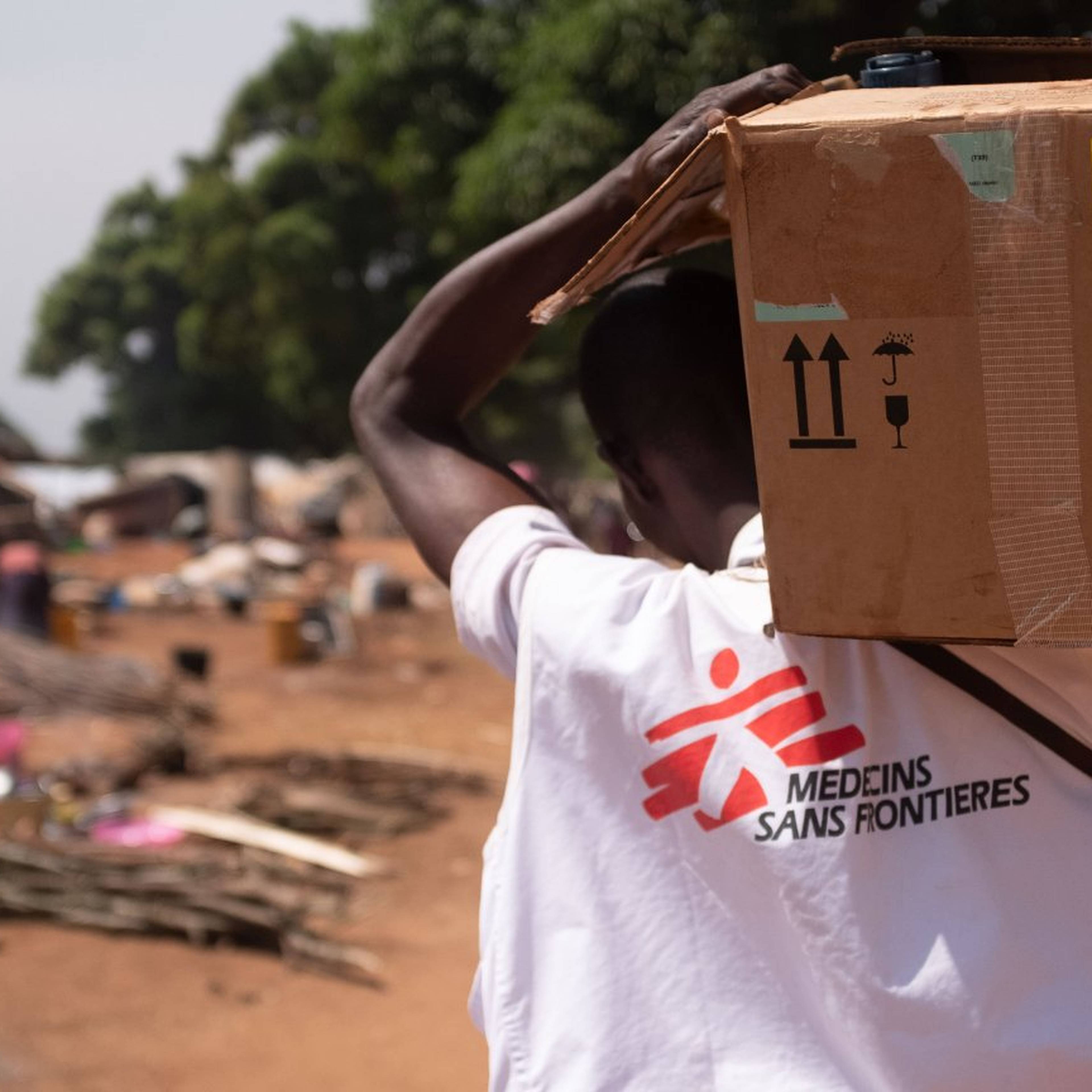 A person wearing a "Médecins Sans Frontières" shirt carries a box in a rural area with tents and trees in the background.