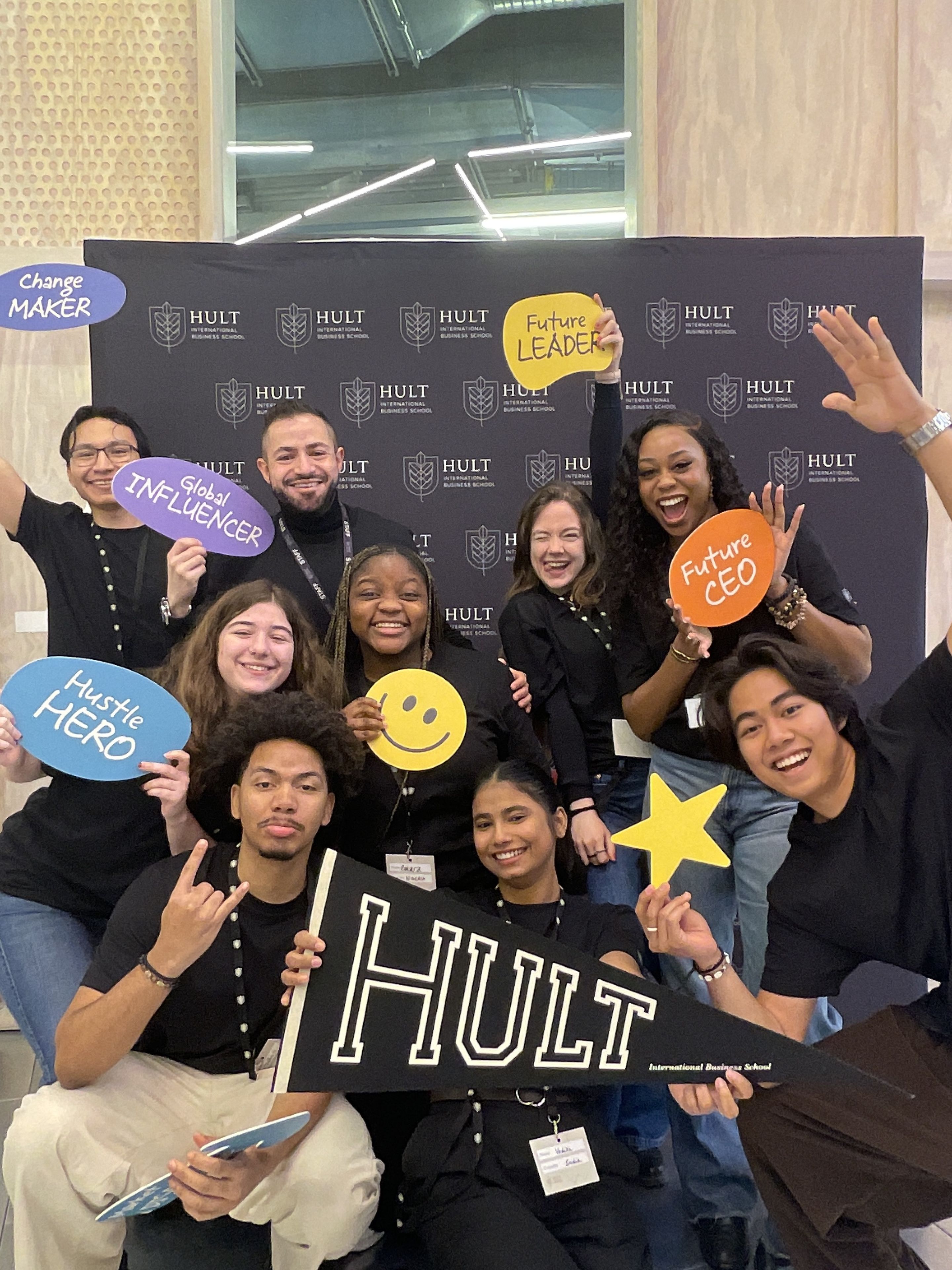 A group of people smiling and holding signs with positive messages, posing in front of a backdrop with the Hult logo.