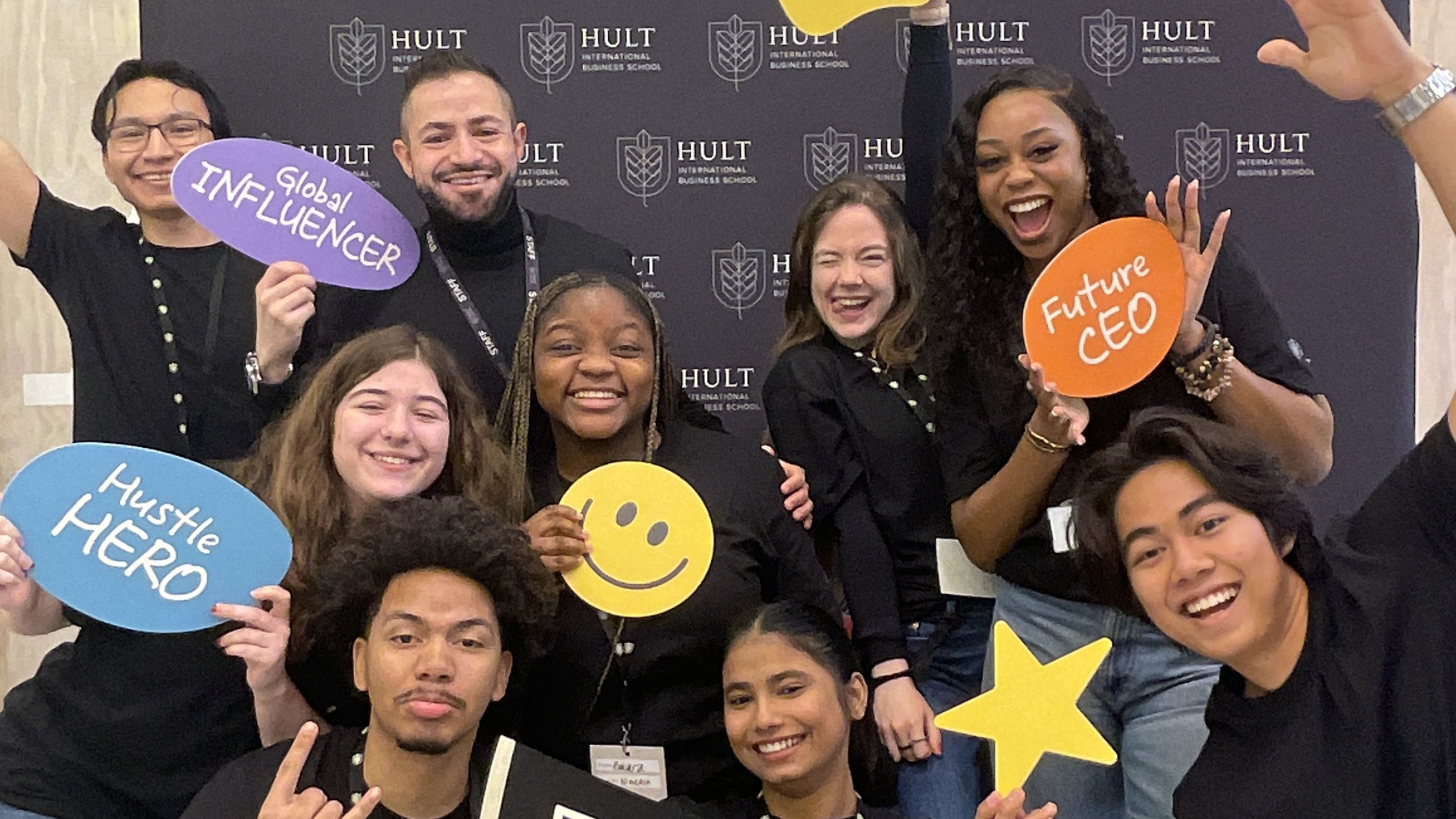 A group of people smiling and holding signs with positive messages, posing in front of a backdrop with the Hult logo.