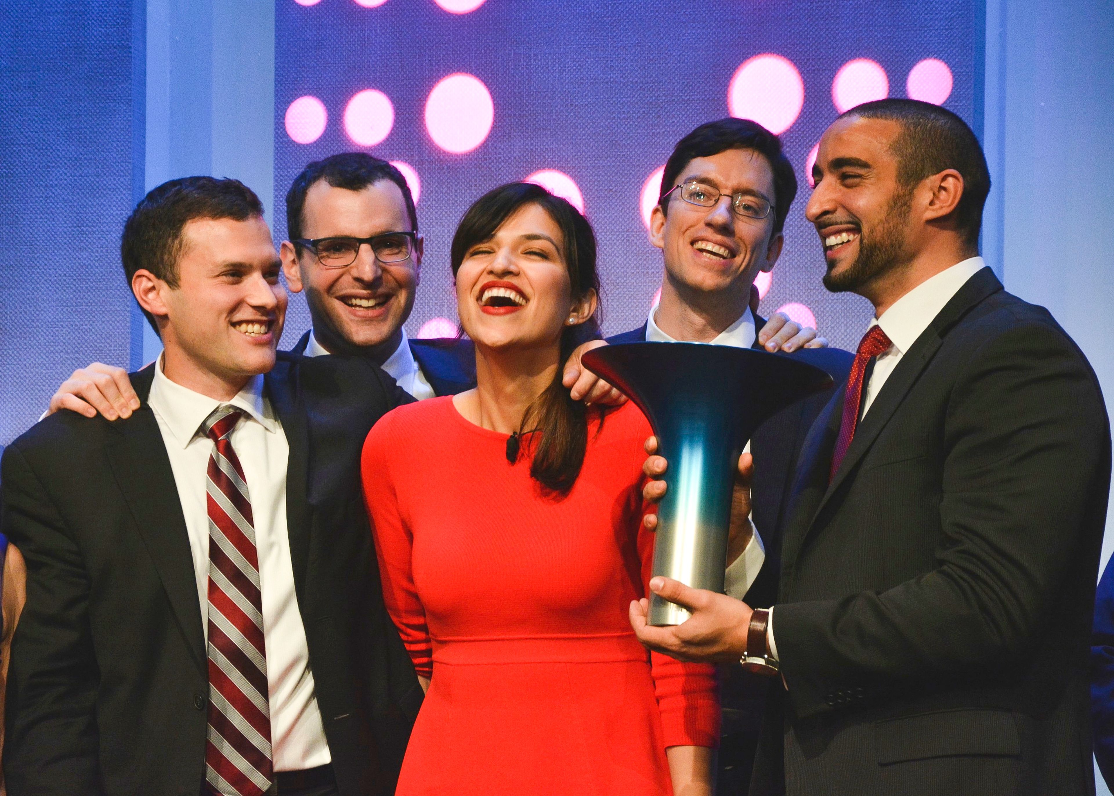 Five people in formal attire smiling on stage, one holding a blue award. Background features pink and purple lights.