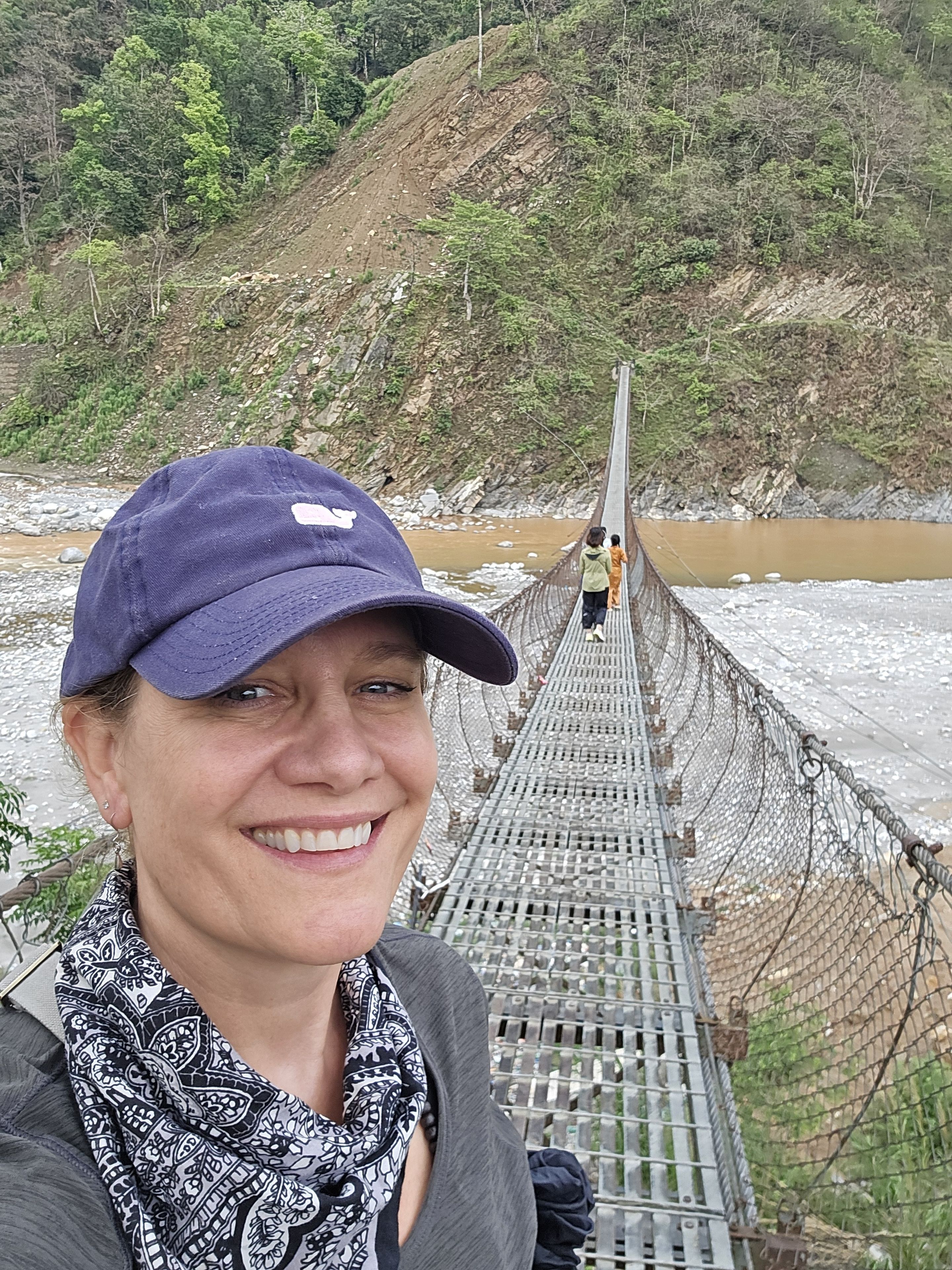 Woman in a cap smiles while standing on a suspension bridge over a river, with hills and trees in the background. Two people walk behind her.