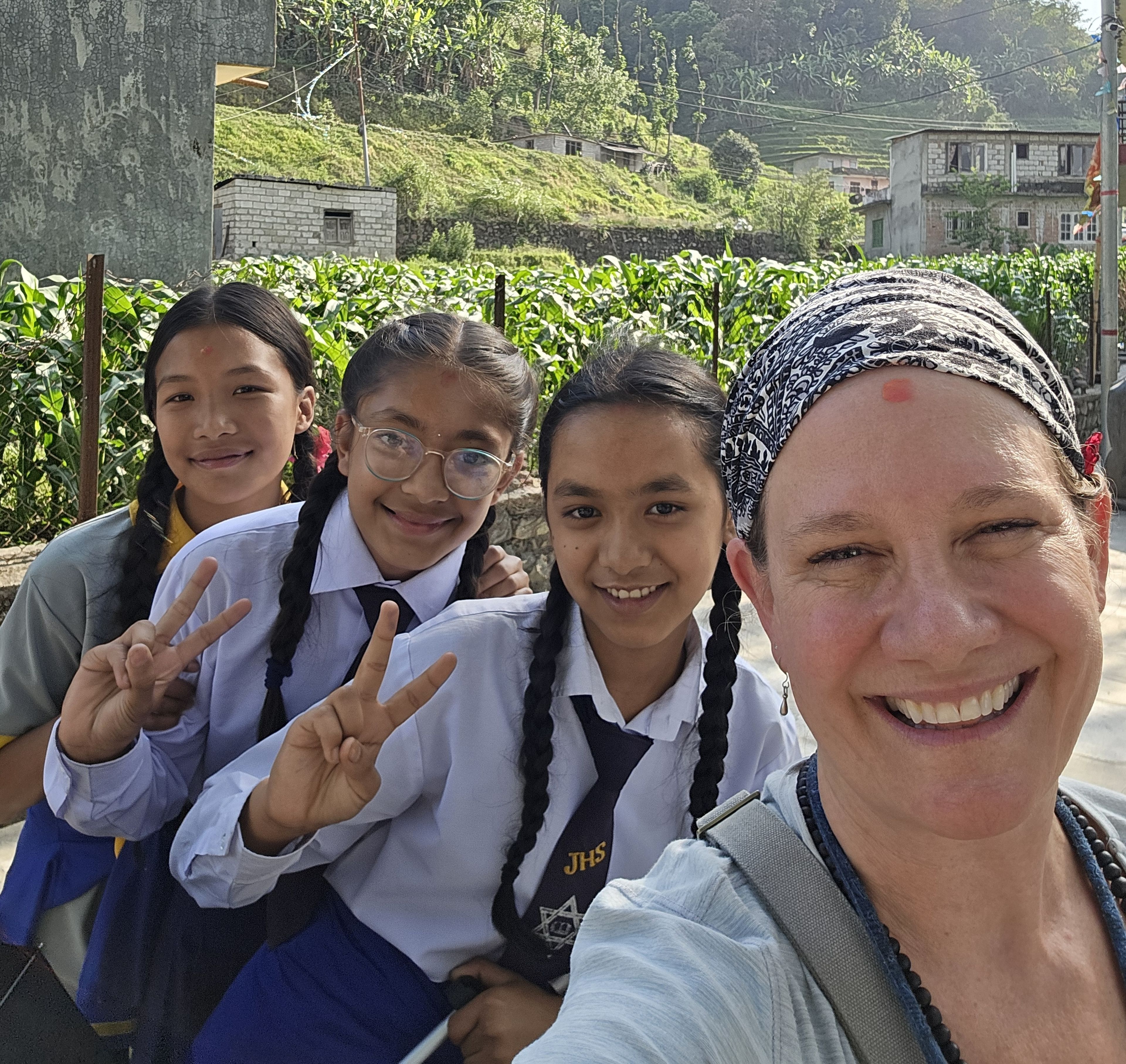 A woman and three schoolgirls smile outdoors. The girls wear uniforms and flash peace signs. Green hills and houses are in the background.