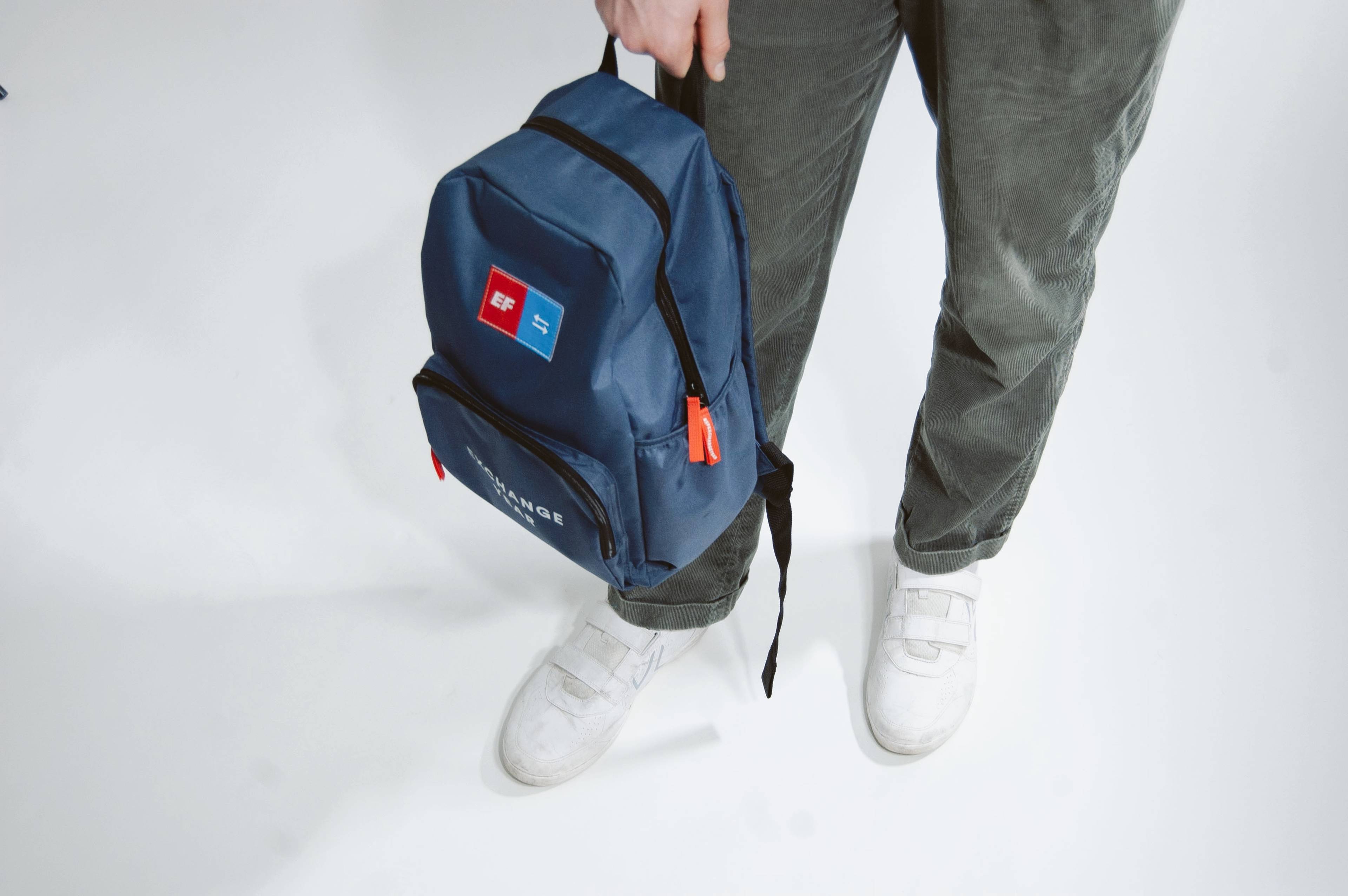 Person holding a blue backpack with red and blue patches, wearing gray pants and white sneakers, standing on a white background.