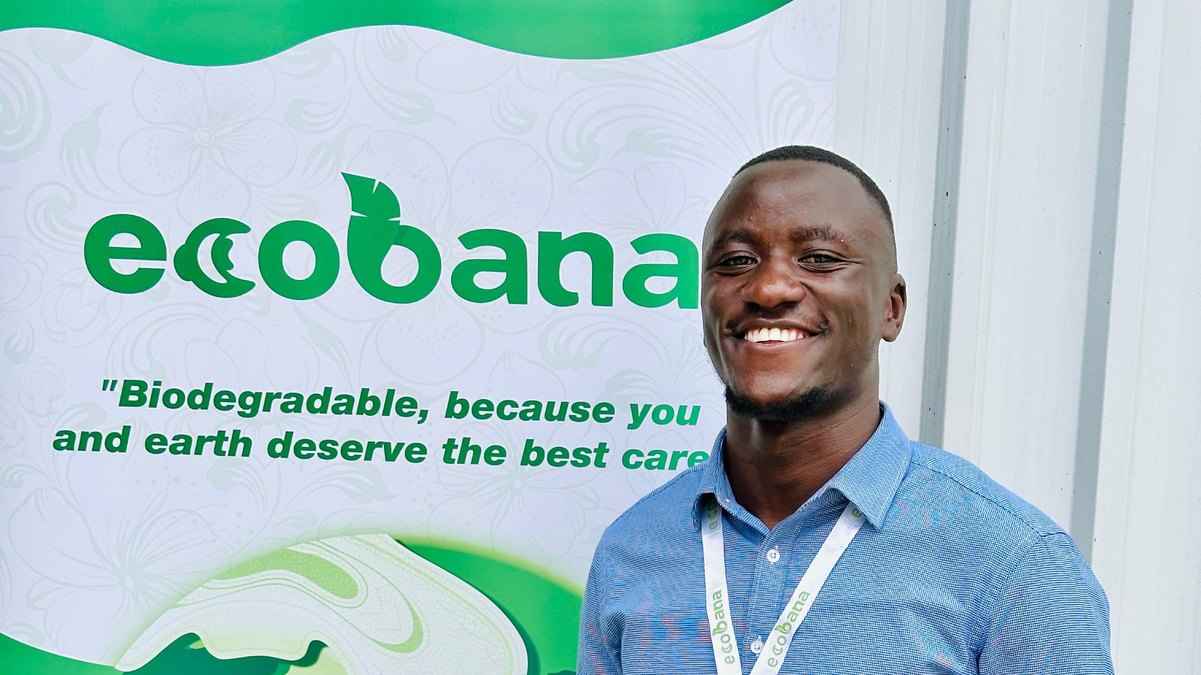 Smiling man in a blue shirt stands in front of an Ecobana banner promoting biodegradable products.