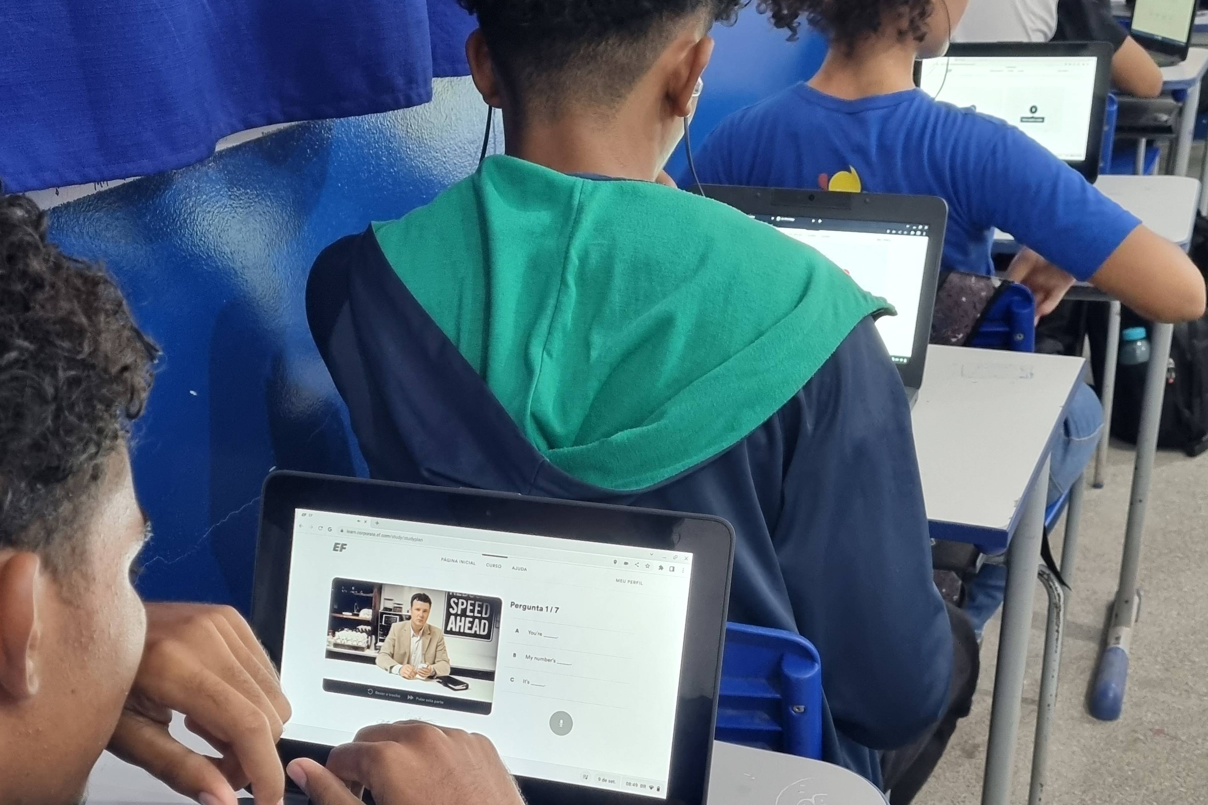 Students sitting in a classroom, using laptops showing EF English Live lessons. Blue curtains and walls are in the background.