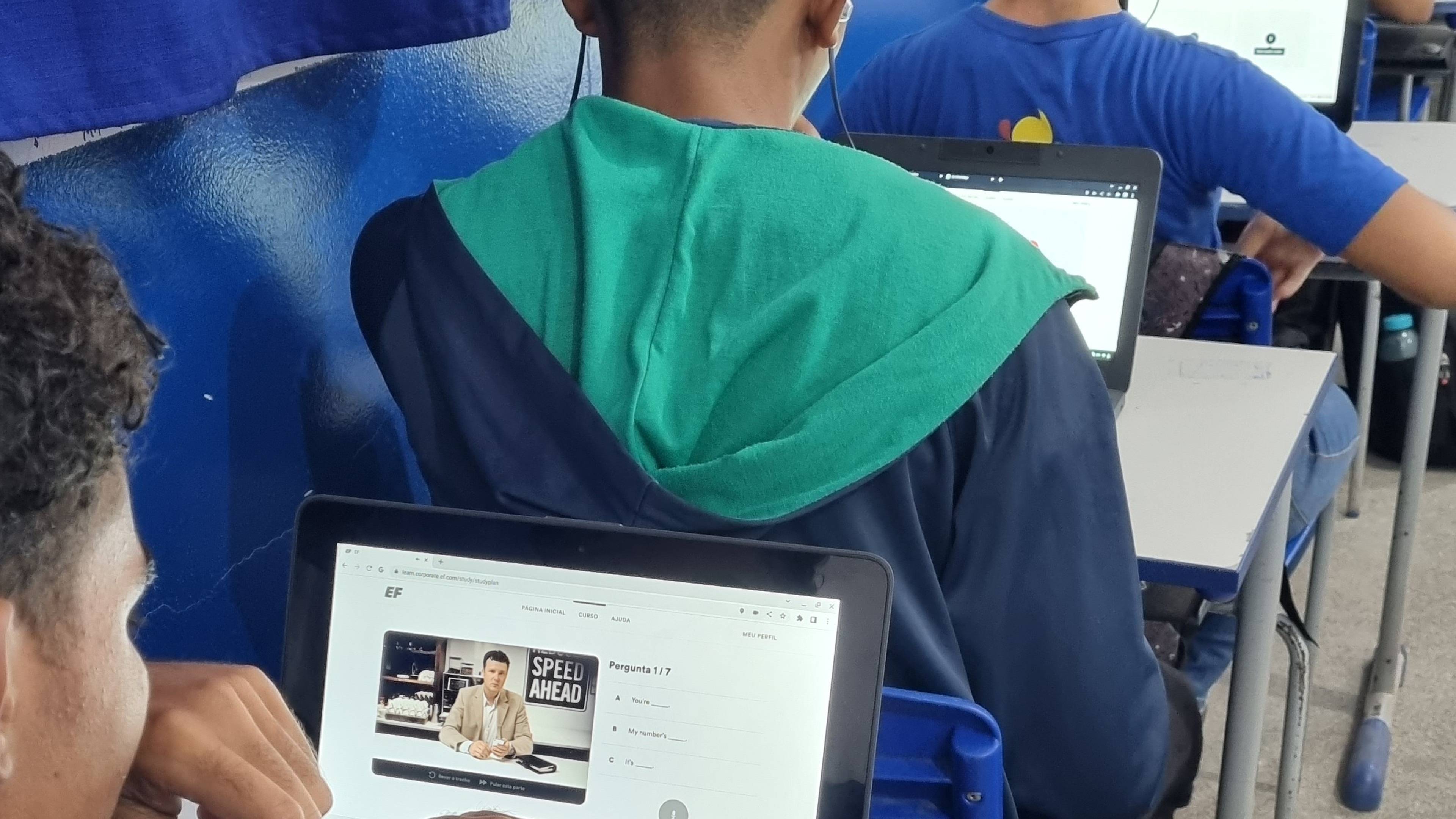 Students sitting in a classroom, using laptops showing EF English Live lessons. Blue curtains and walls are in the background.