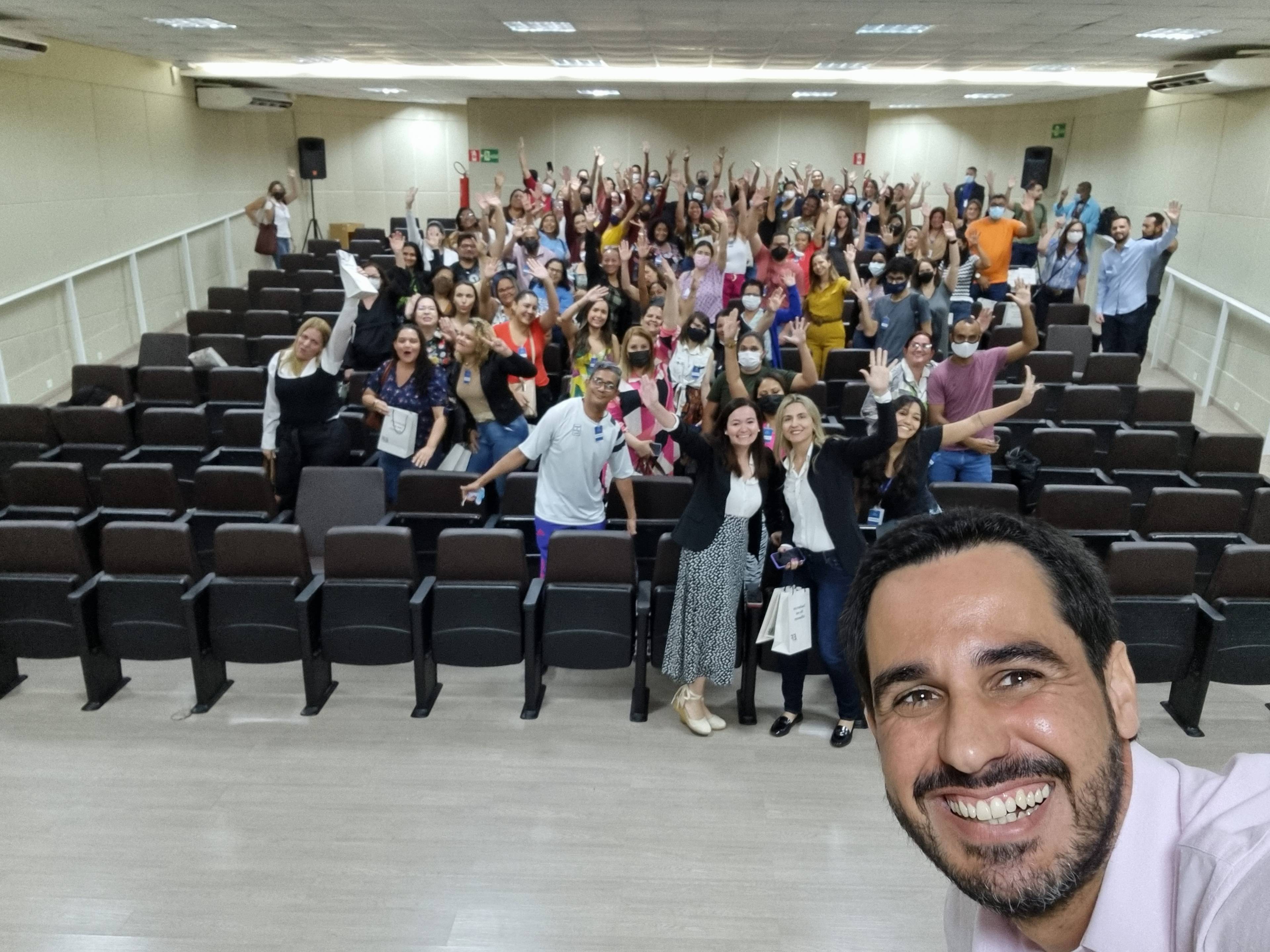 A man takes a selfie in a lecture hall filled with cheerful teachers, many of whom are waving and smiling at the camera.