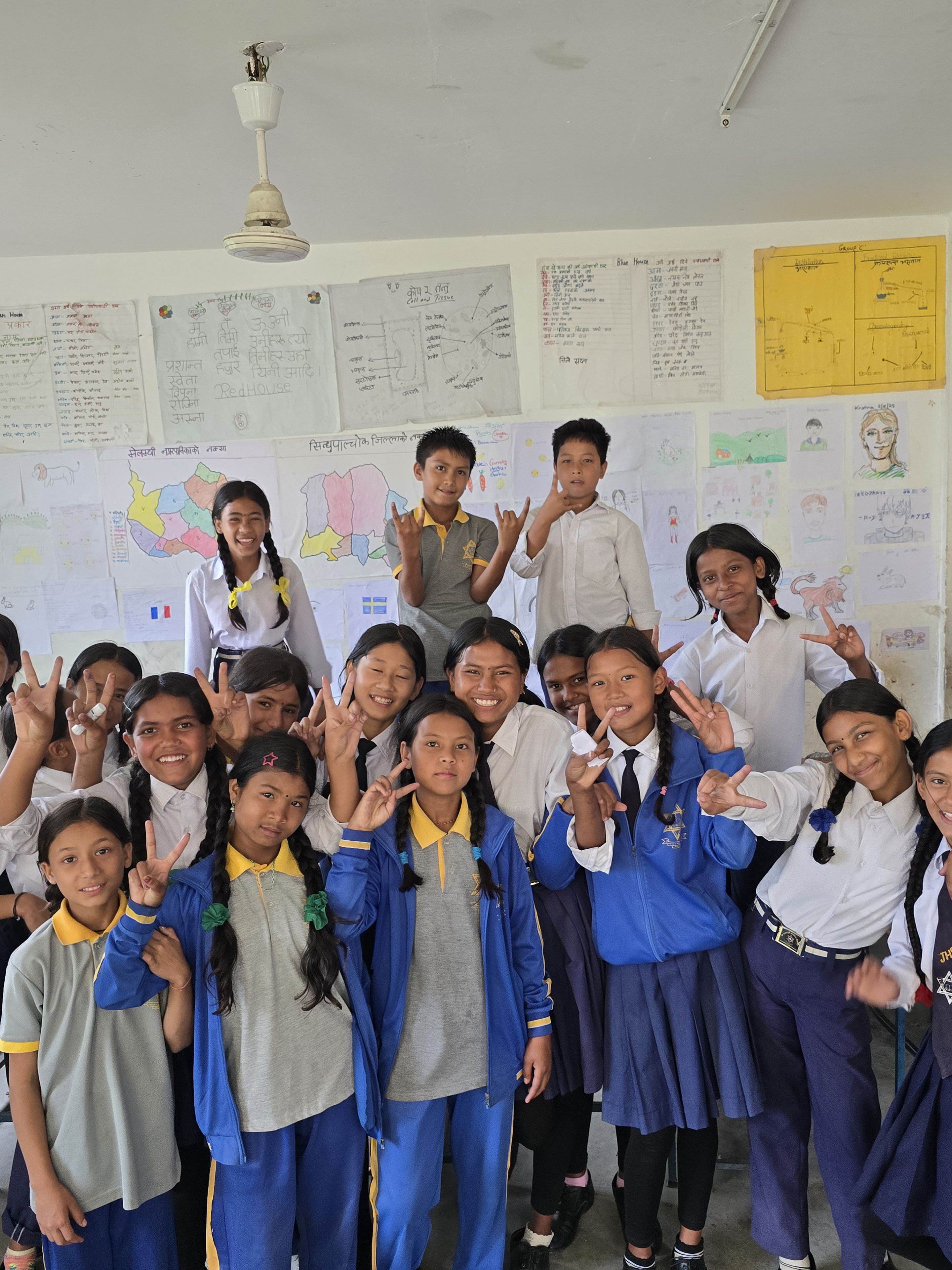A group of students in school uniforms pose cheerfully in a classroom, with colorful posters on the walls behind them.