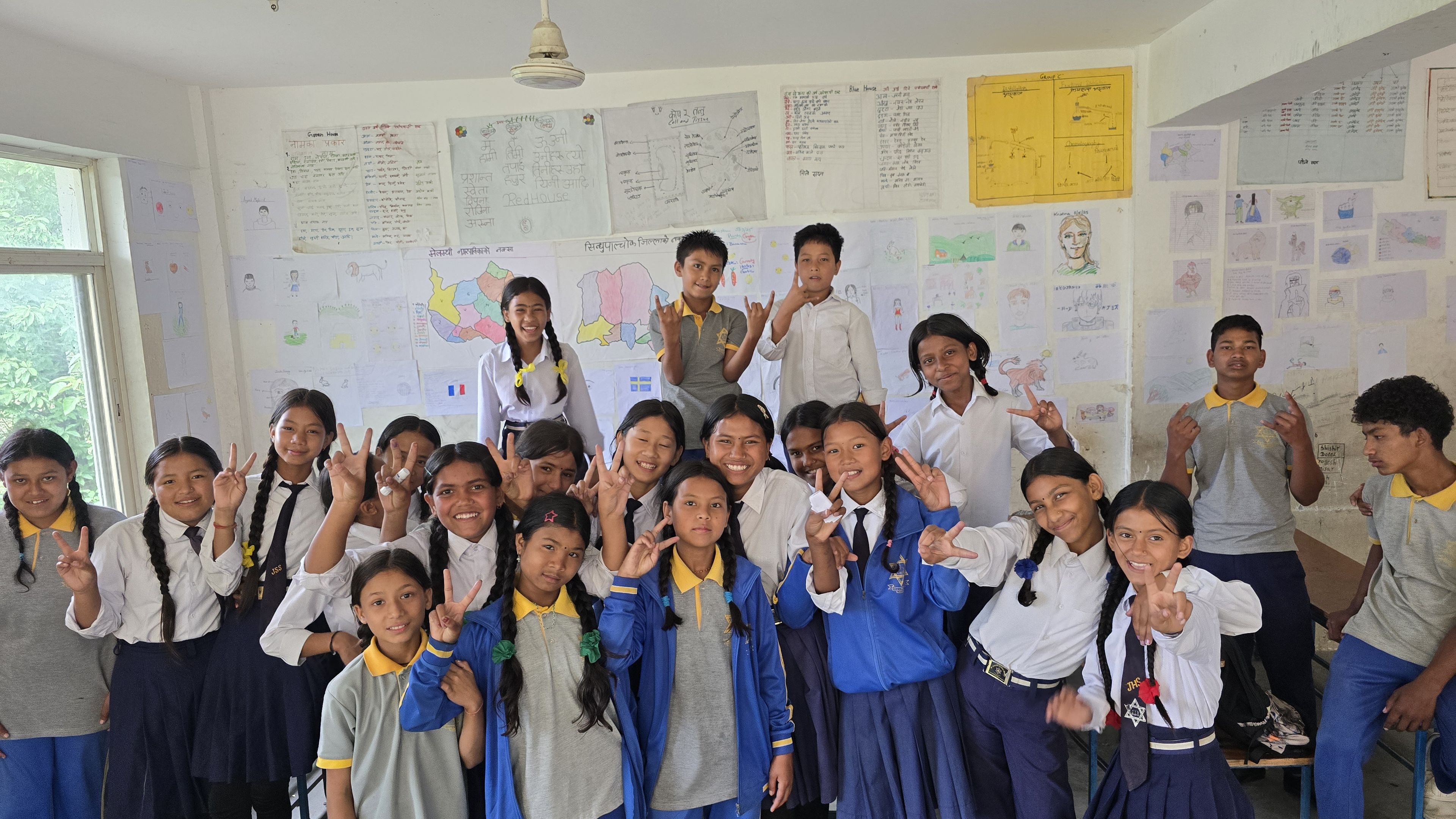 A group of students in school uniforms pose cheerfully in a classroom, with colorful posters on the walls behind them.