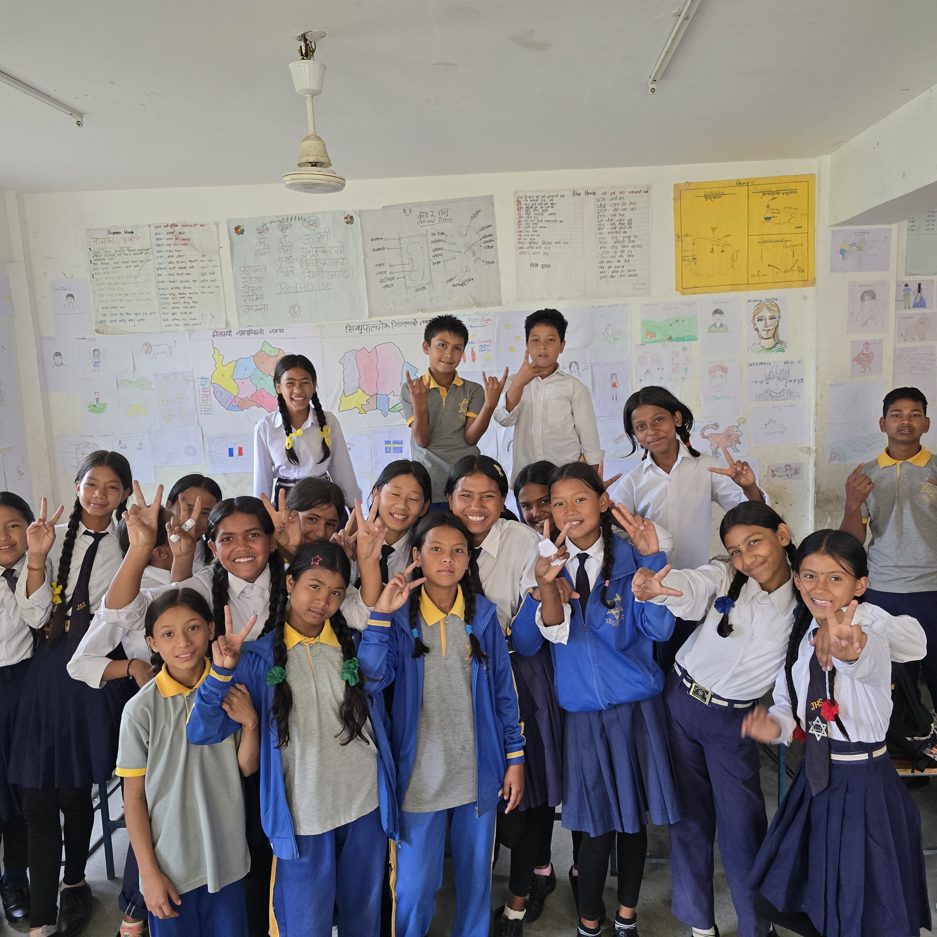 A group of students in school uniforms pose cheerfully in a classroom, with colorful posters on the walls behind them.
