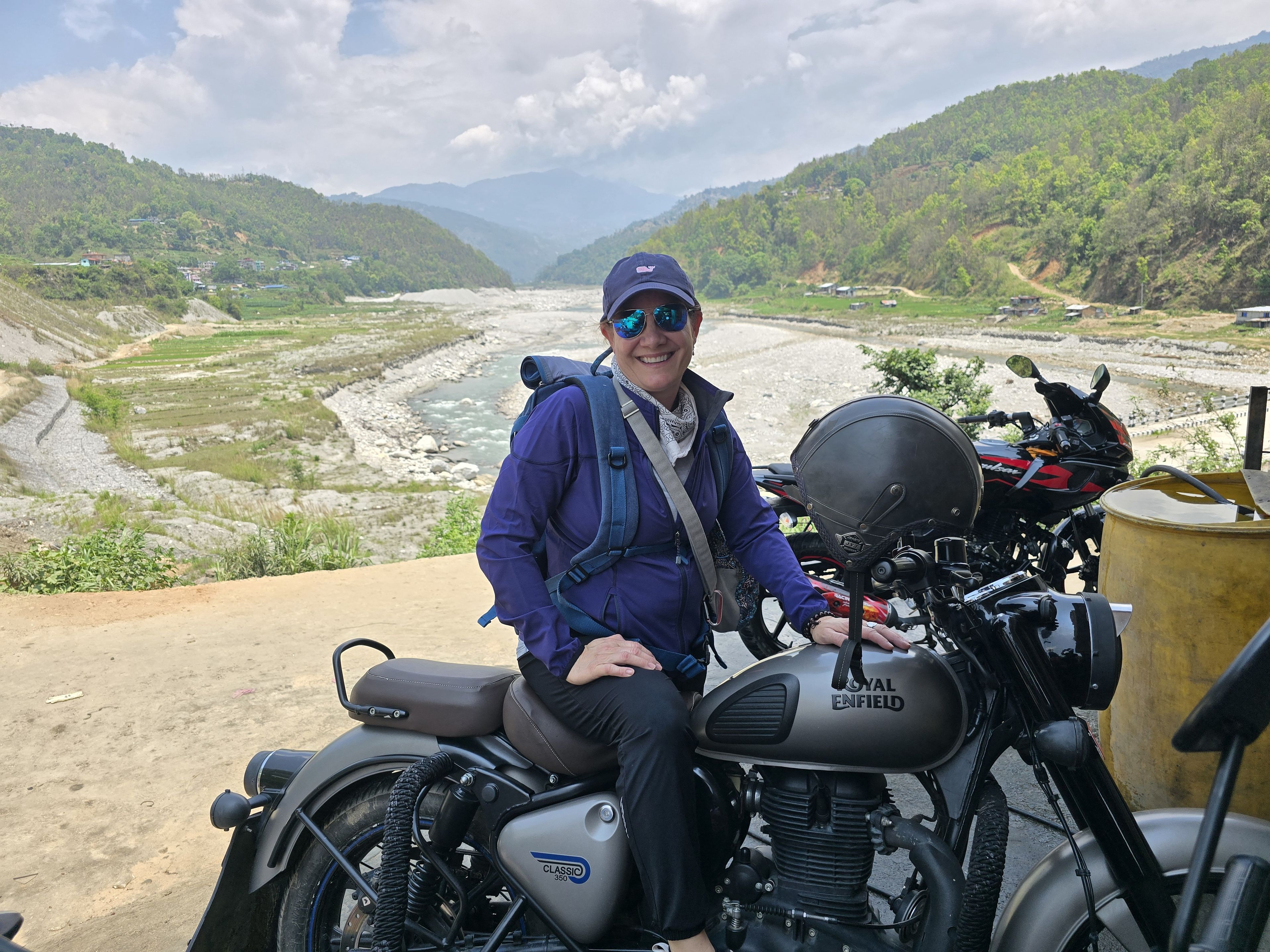 Woman in outdoor gear sits on a Royal Enfield motorcycle, with a scenic river and mountain backdrop under a partly cloudy sky.