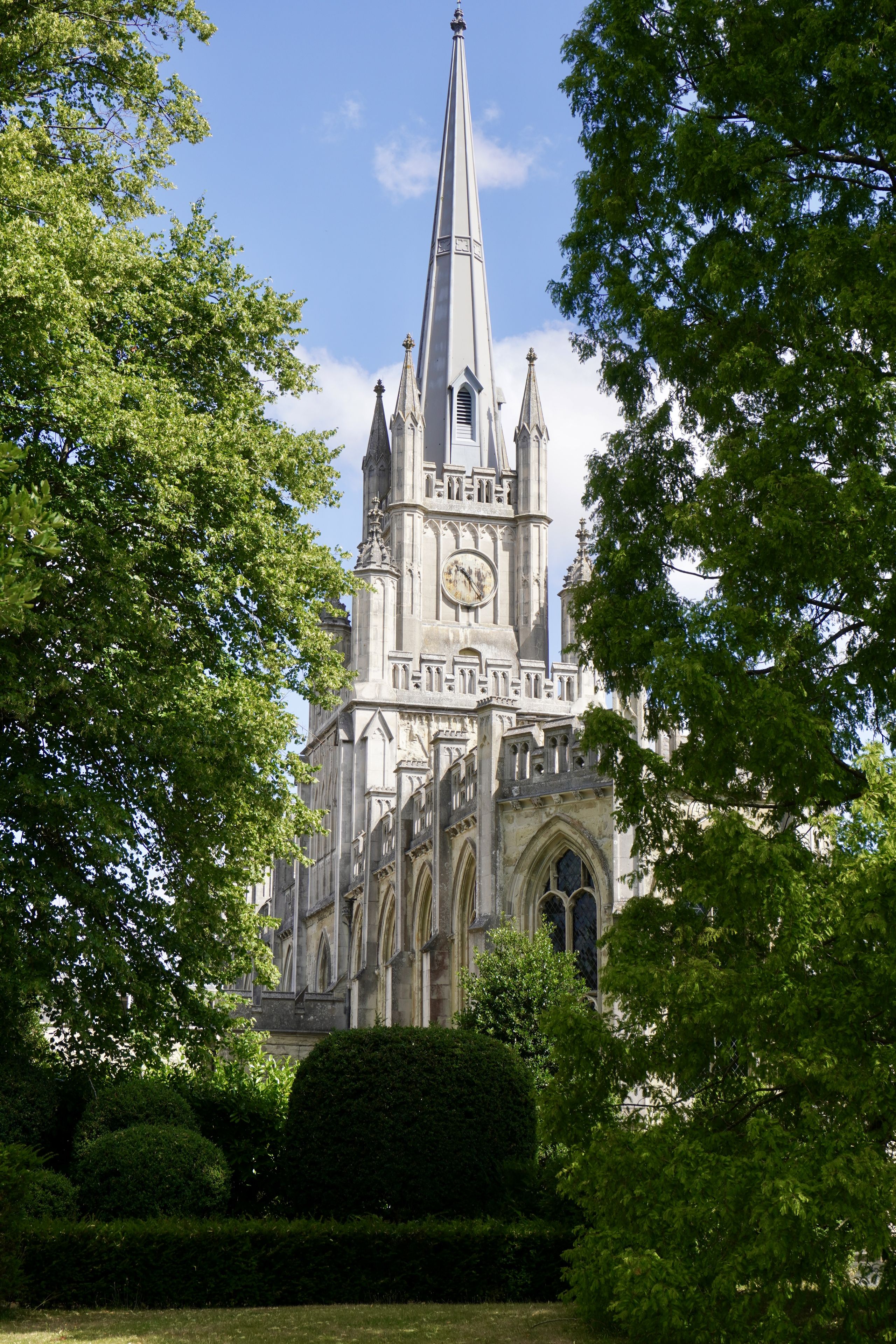 A Gothic-style church with a tall spire and clock is framed by lush green trees under a blue sky.