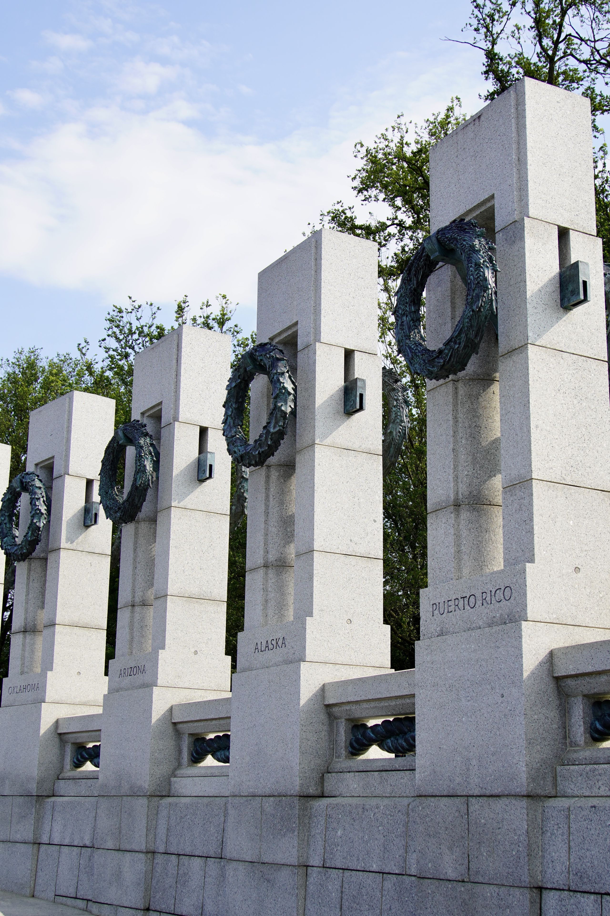 Stone columns with wreaths, each inscribed with names: Oklahoma, Arizona, Alaska, Puerto Rico, against a backdrop of trees and sky.