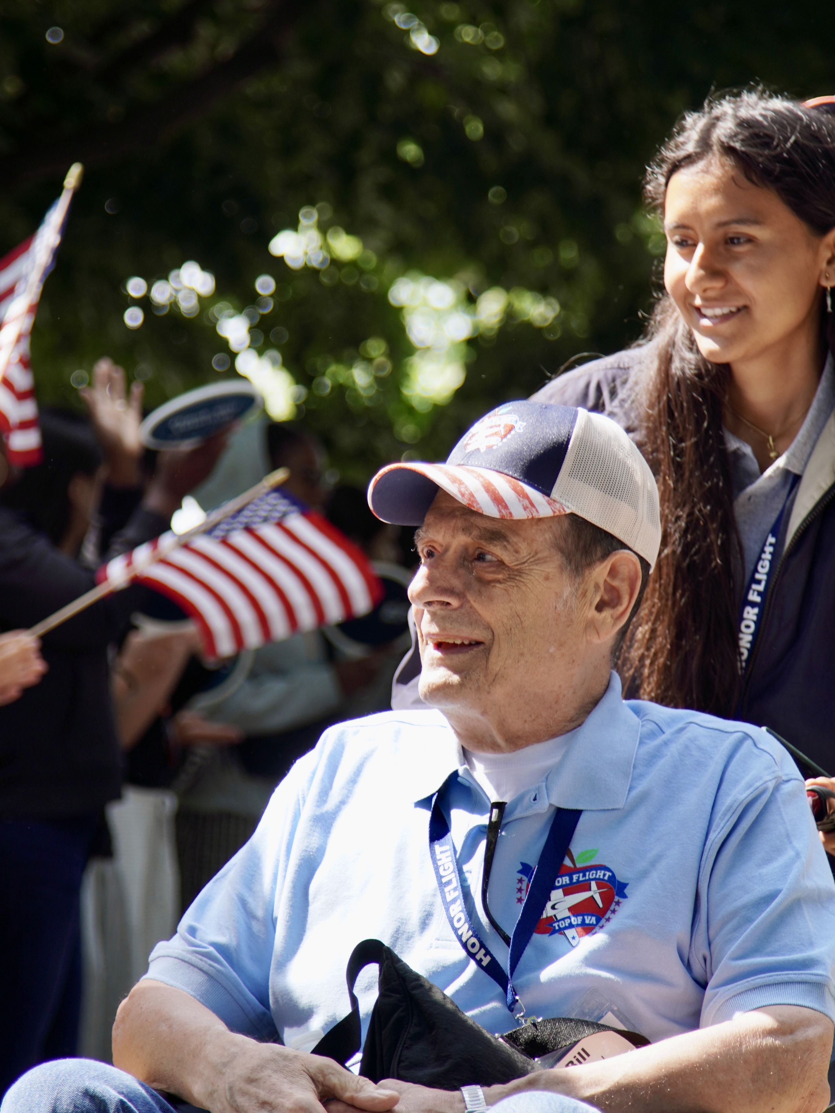 A smiling older man in a wheelchair is pushed by a young woman. They are outdoors, surrounded by people waving American flags.