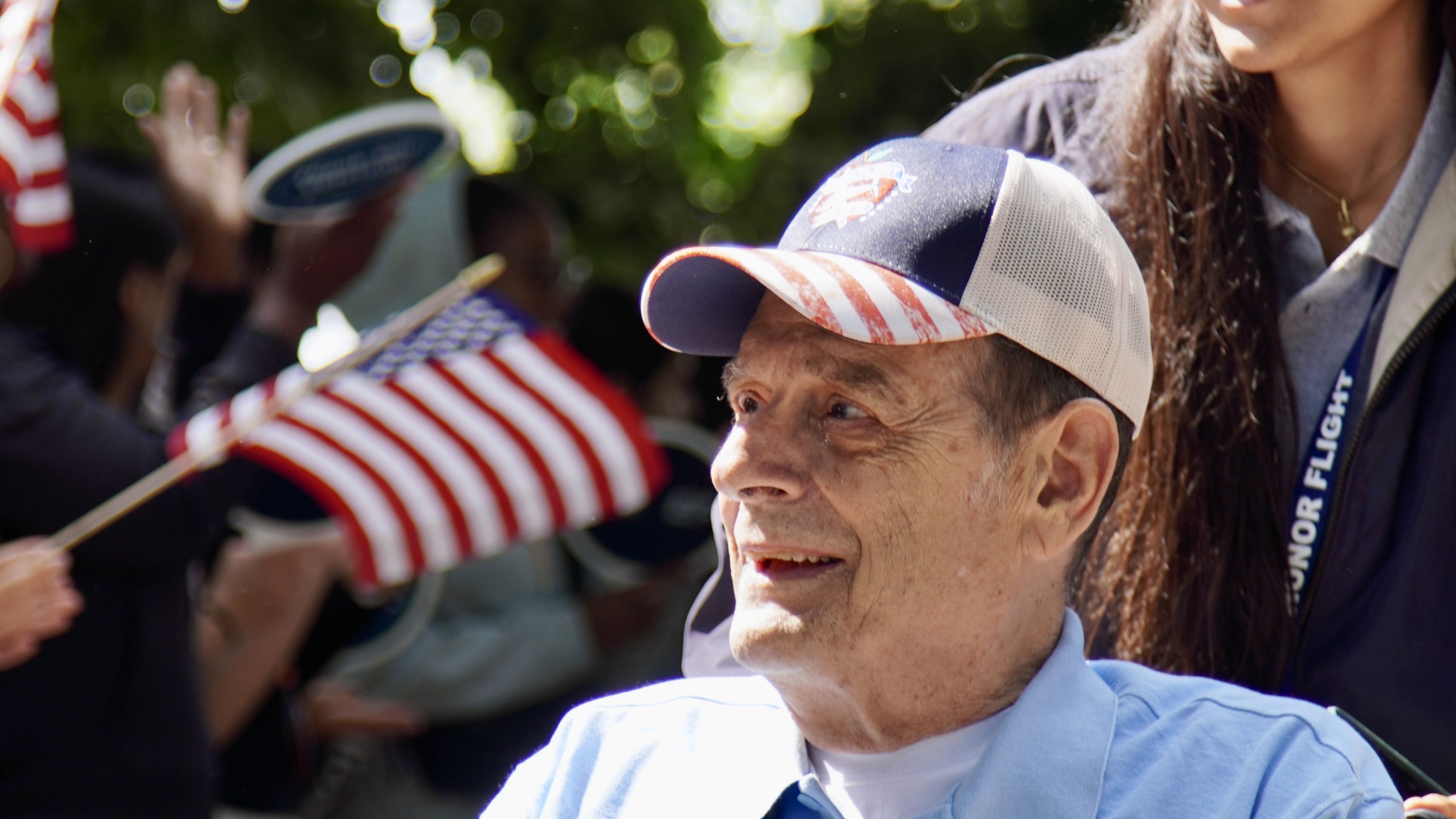 A smiling older man in a wheelchair is pushed by a young woman. They are outdoors, surrounded by people waving American flags.