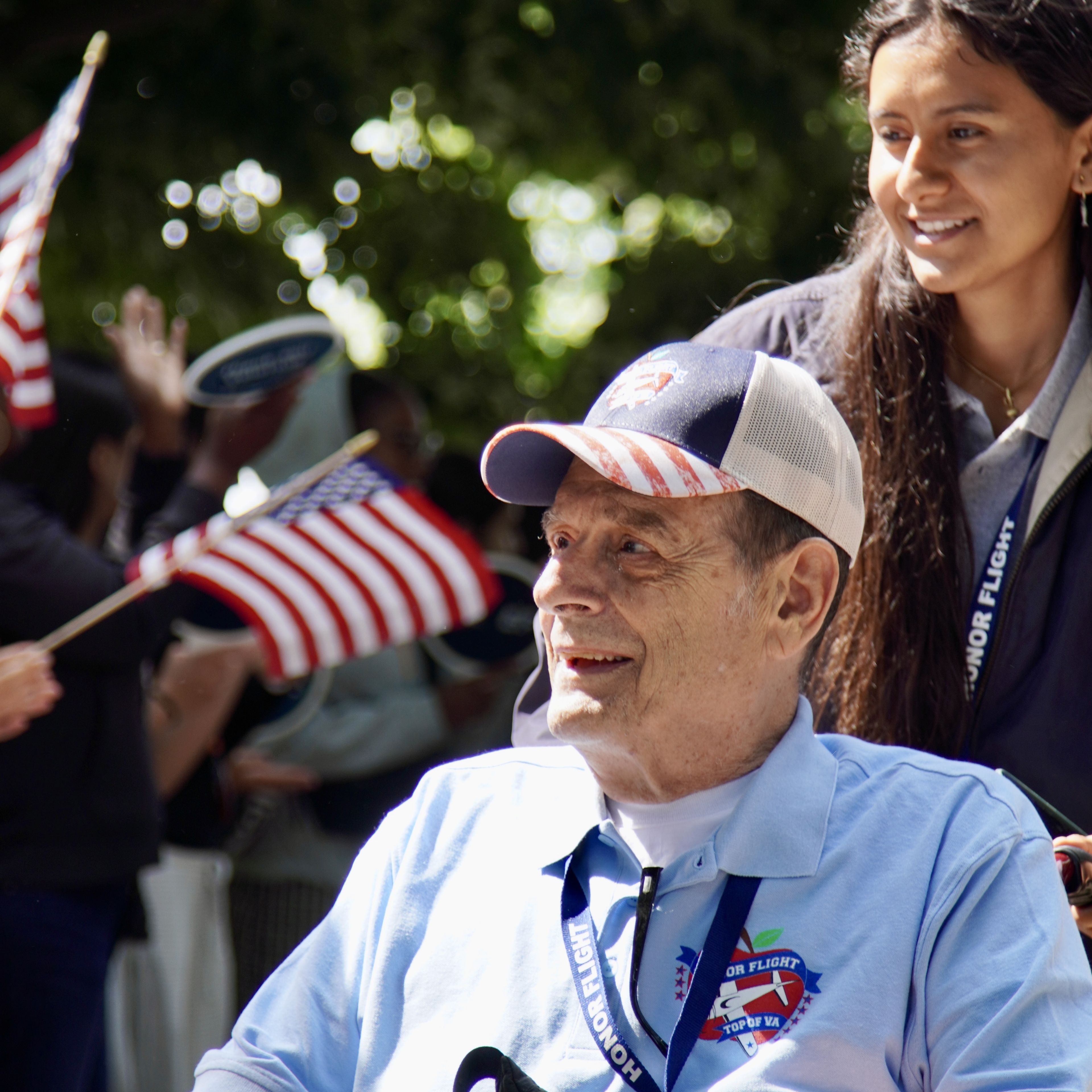 A smiling older man in a wheelchair is pushed by a young woman. They are outdoors, surrounded by people waving American flags.