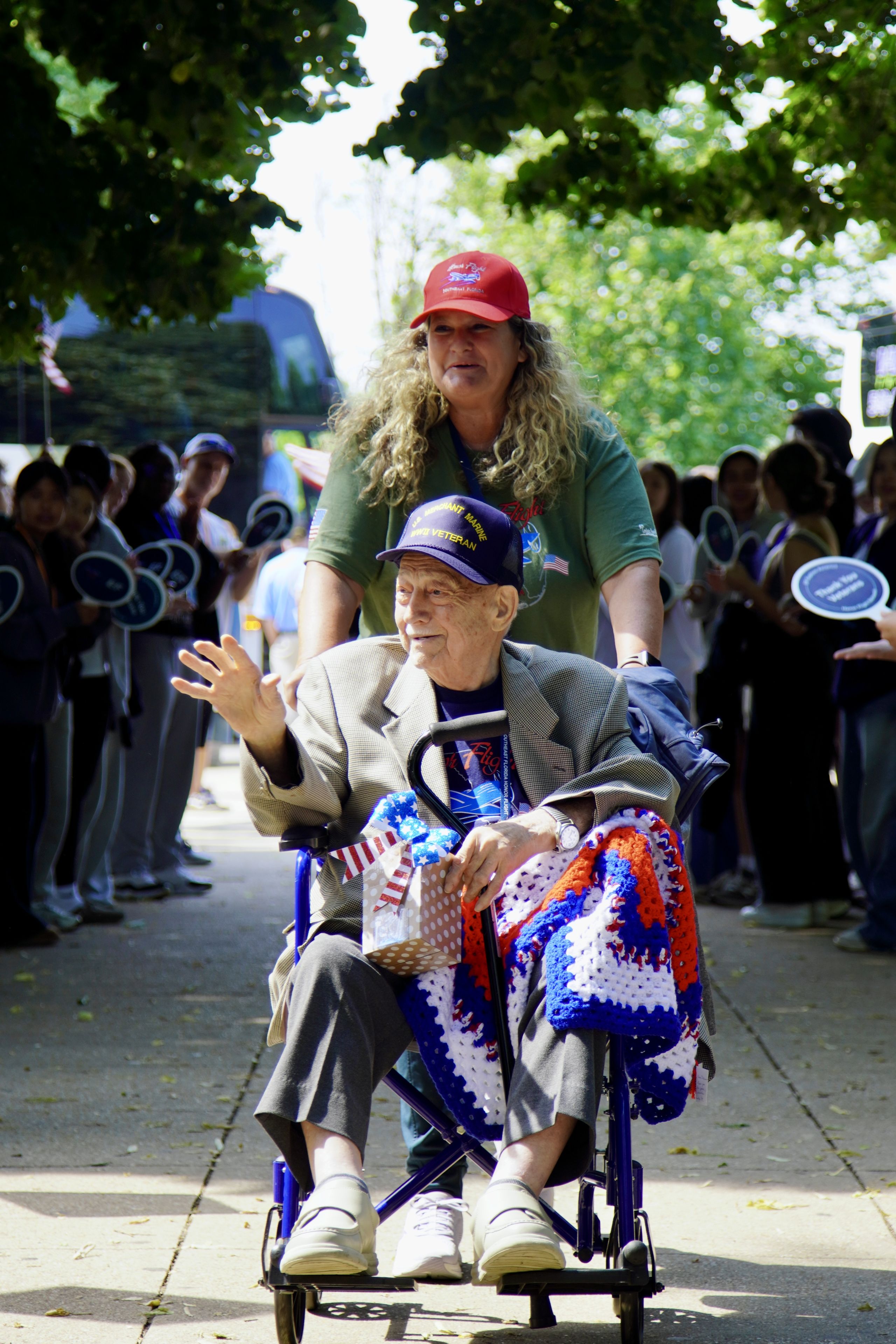 Joe in a wheelchair, wearing a cap and waving, is pushed by a woman. A crowd claps in the background.