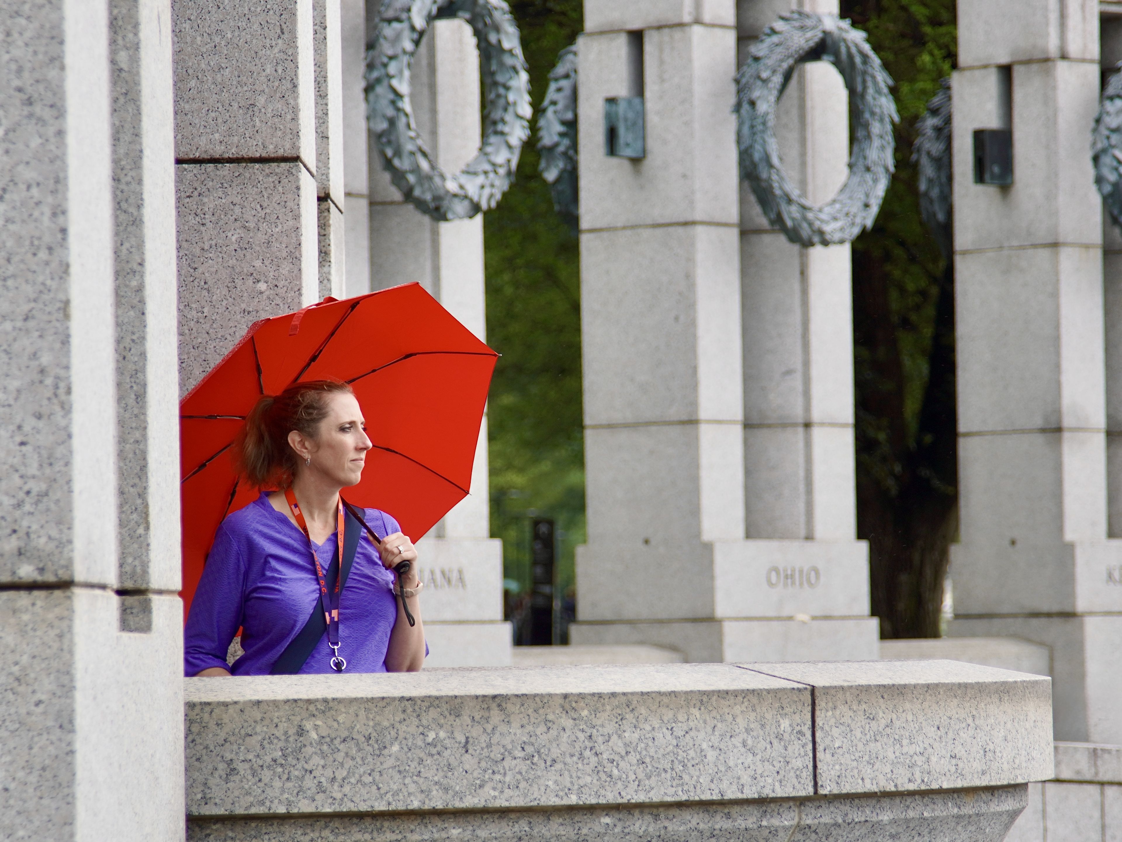 Woman with red umbrella stands near stone columns, wearing a purple top, looking into the distance. Wreaths adorn the columns.