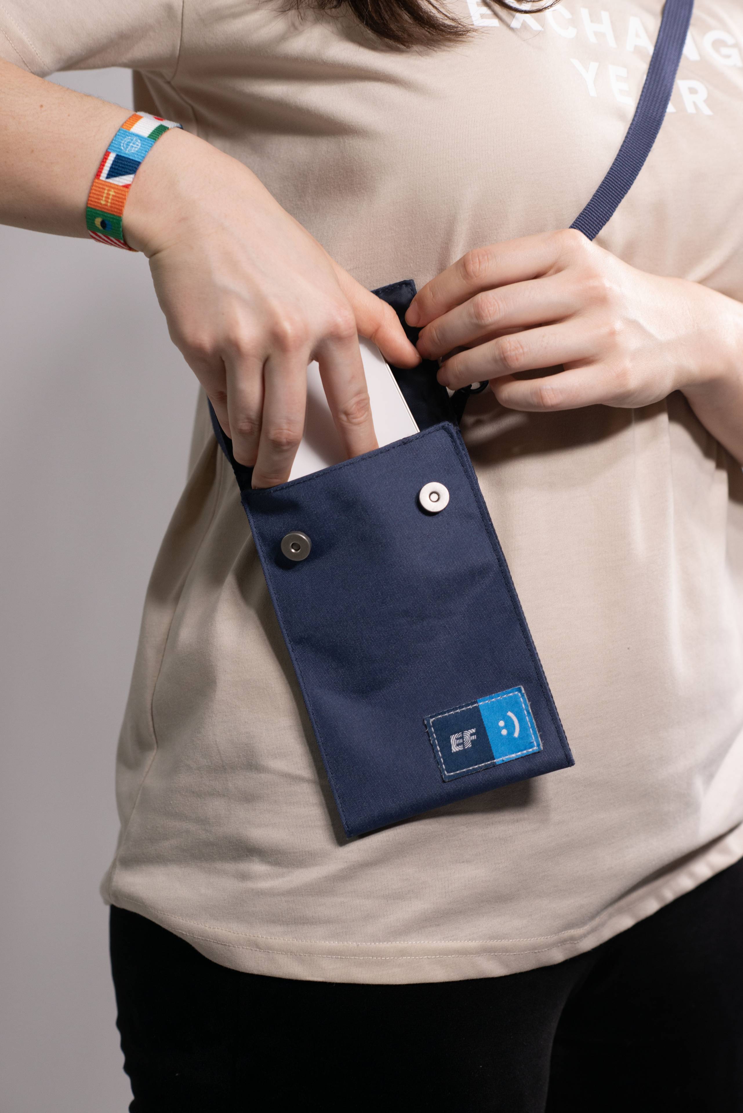 Person placing a white object into a small blue shoulder bag with a logo, wearing a beige shirt and colorful wristband. The bag has the EF logo