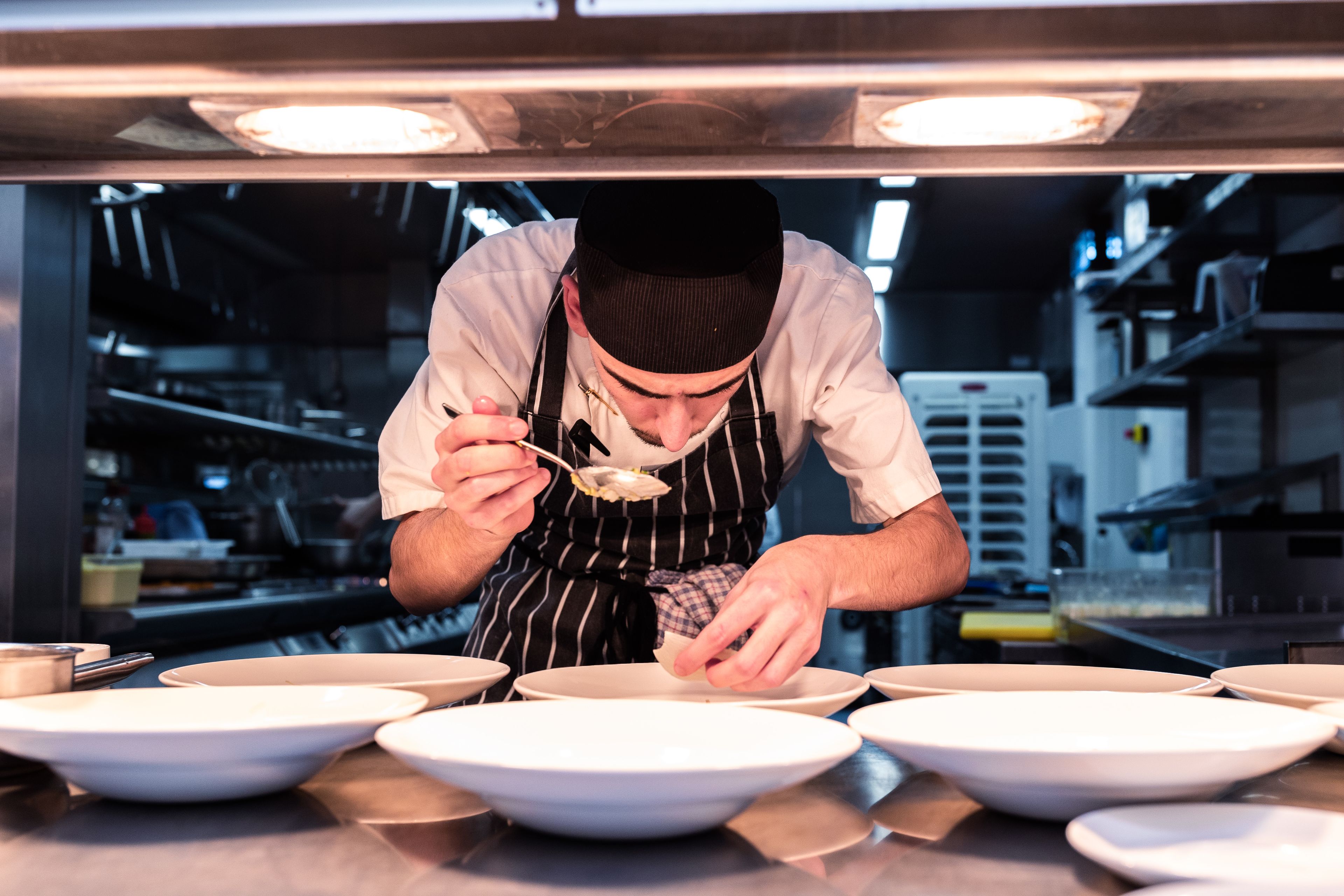 Chef in a striped apron carefully plating dishes in a professional kitchen, focusing intently under warm lighting.