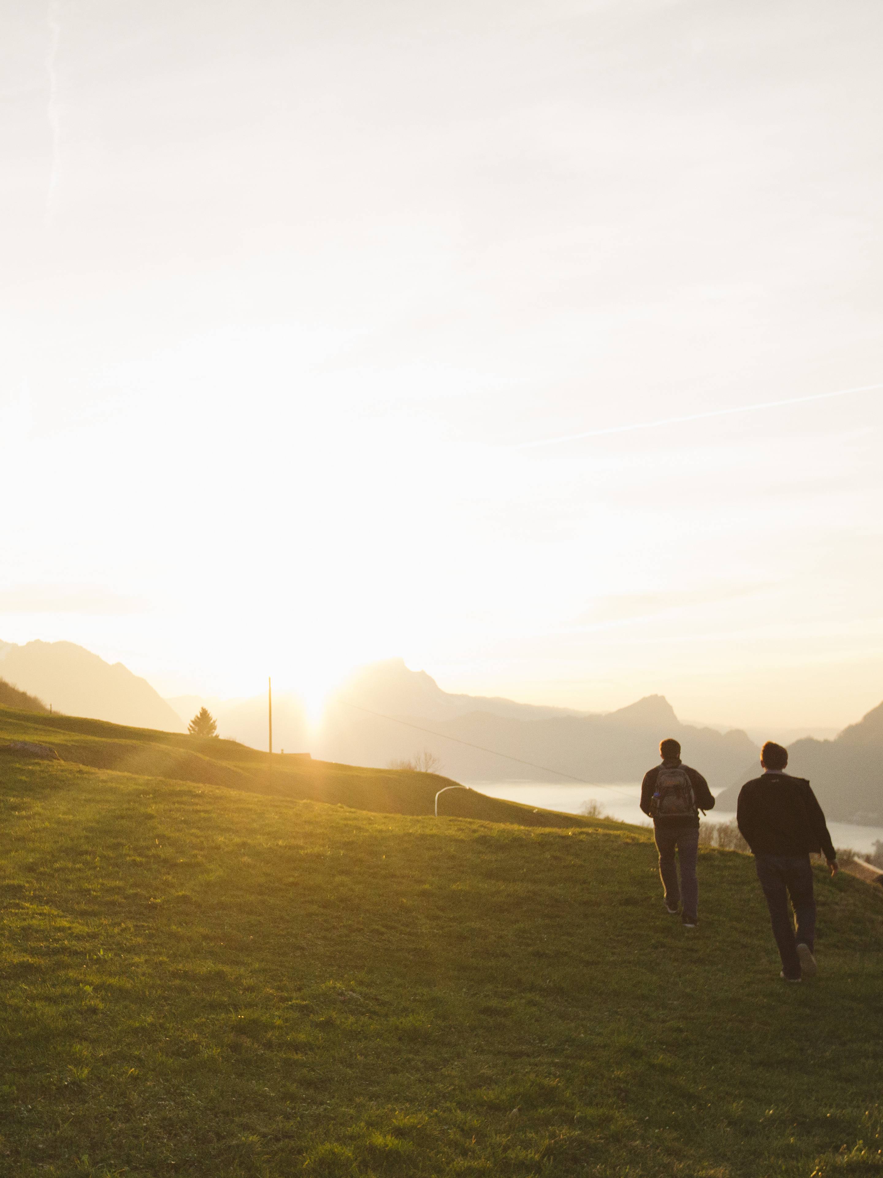 Two people walk across a grassy field at sunset, surrounded by mountains and a distant lake, with a warm, golden sky.