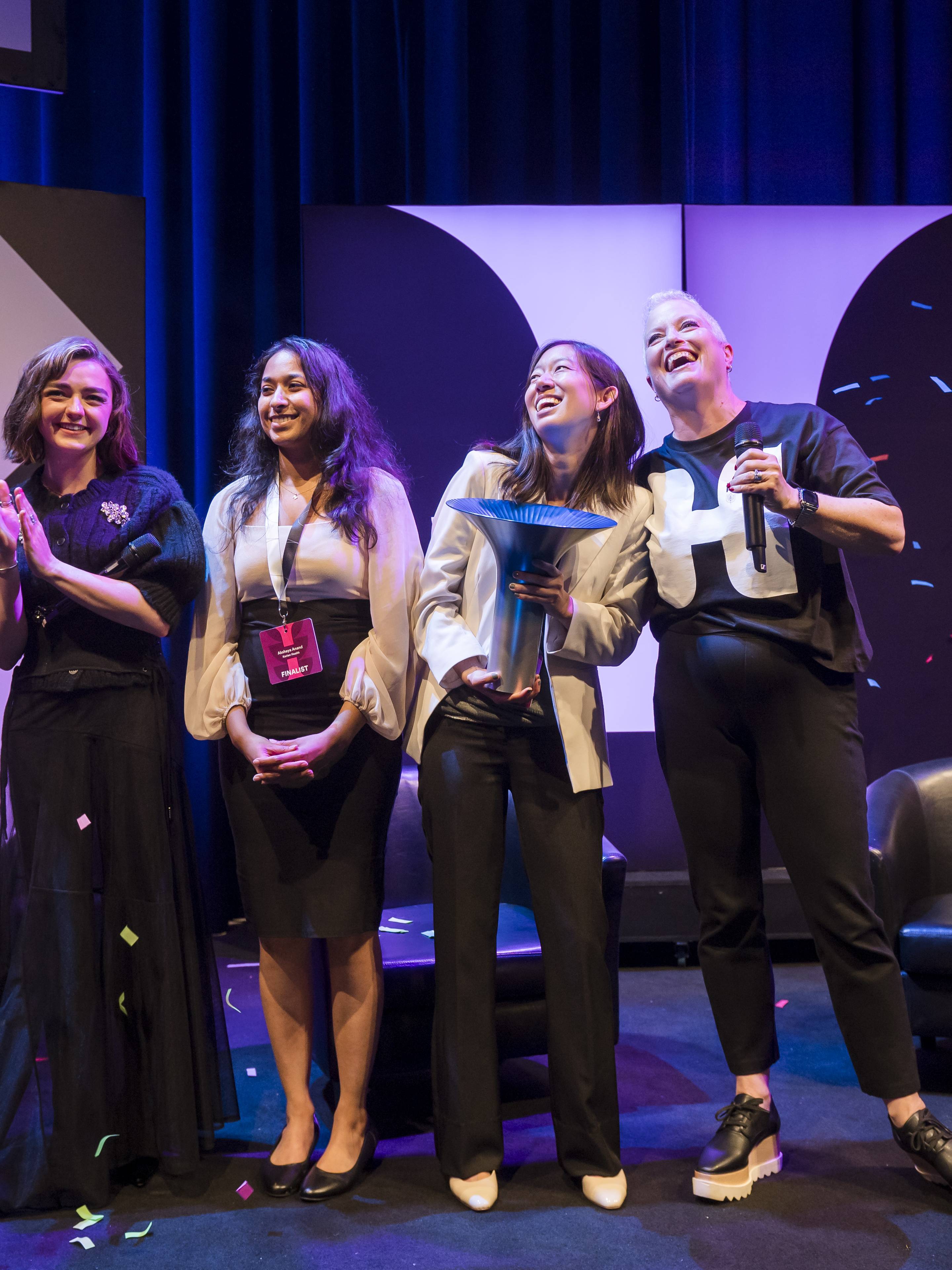 Four women on stage celebrate with confetti falling, two holding trophies, against a backdrop with geometric patterns.