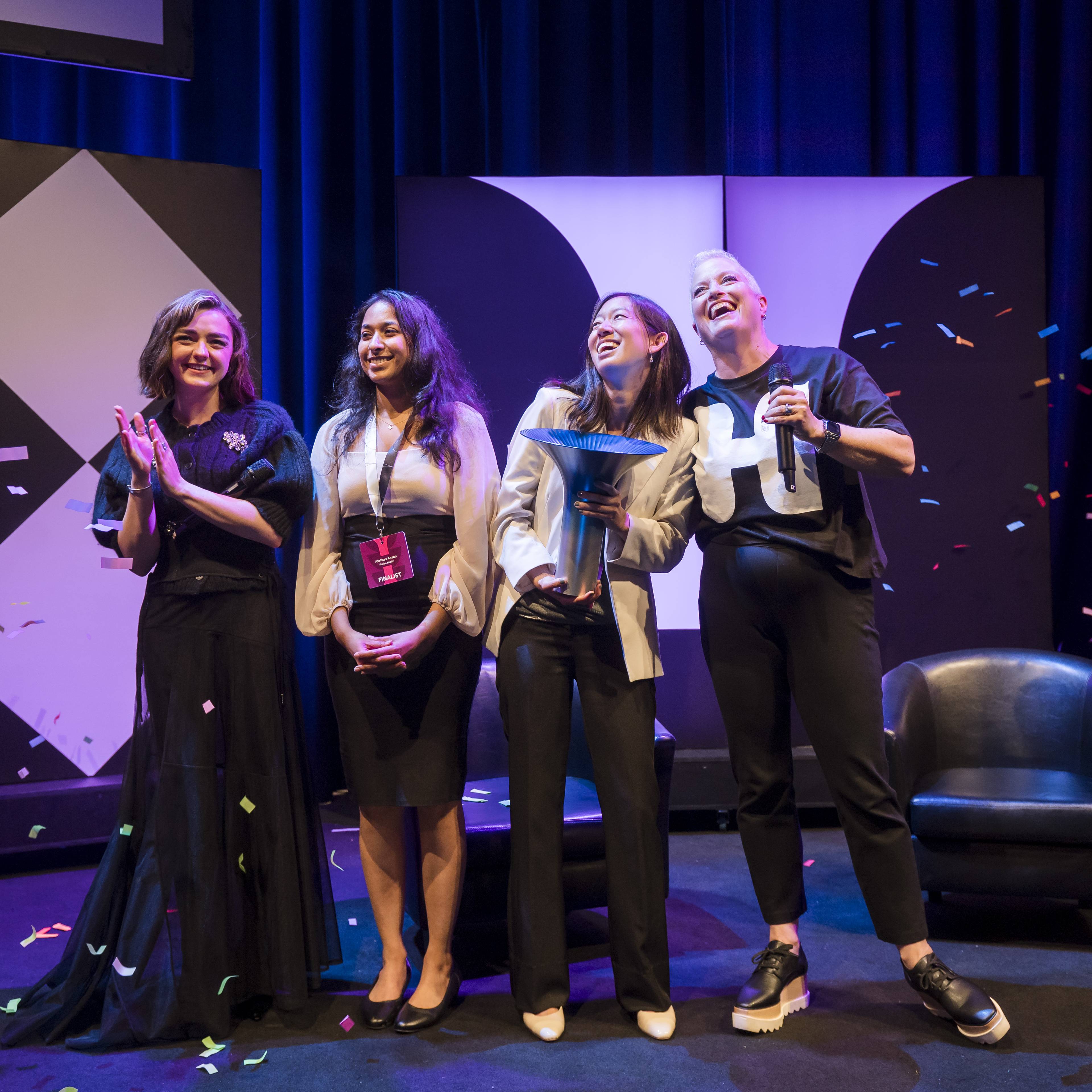 Four women on stage celebrate with confetti falling, two holding trophies, against a backdrop with geometric patterns.