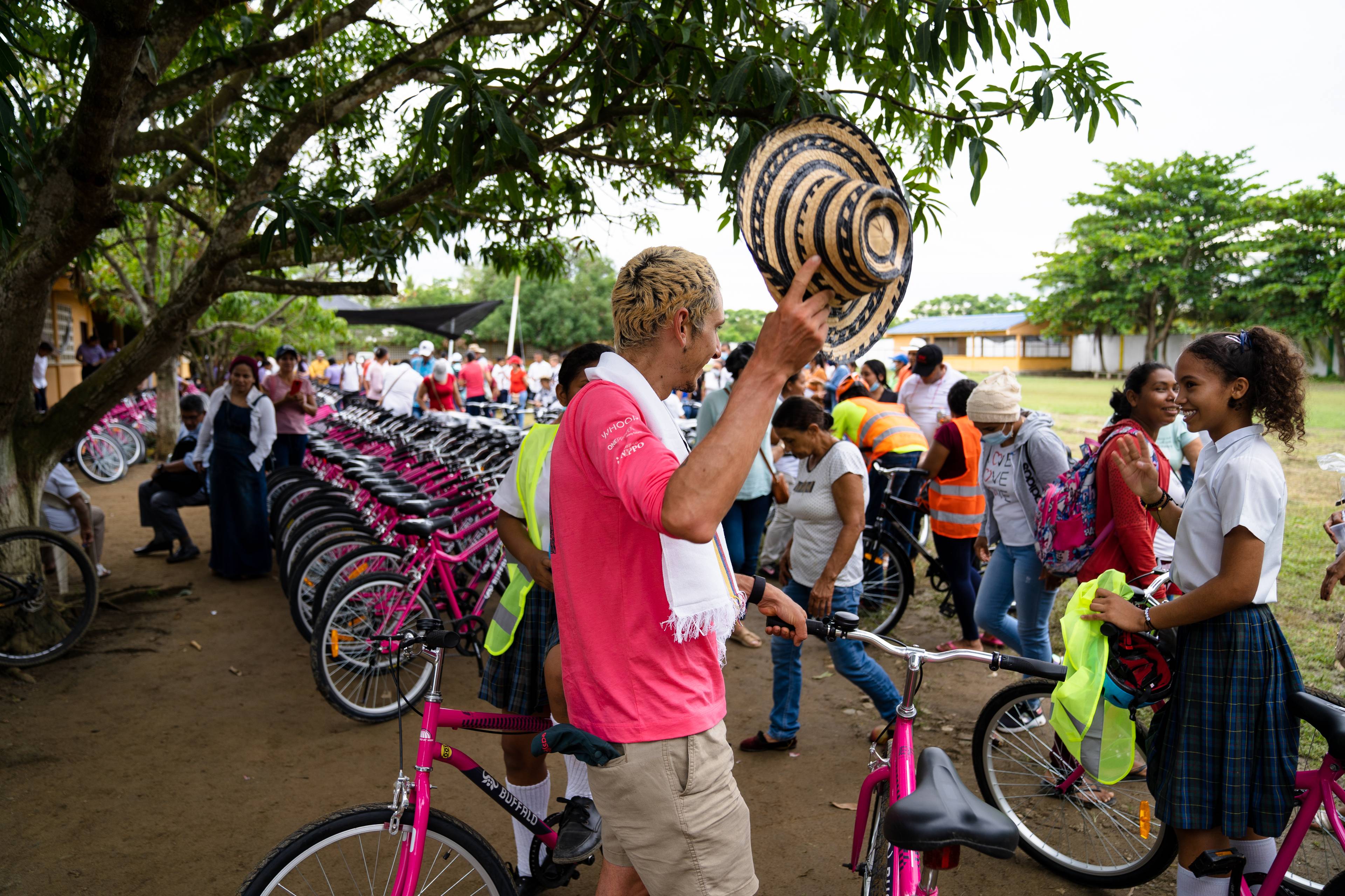 A group of people gather outdoors near a row of pink bicycles. A man (Lachlan) in a pink shirt holds up a hat, and others are interacting nearby.