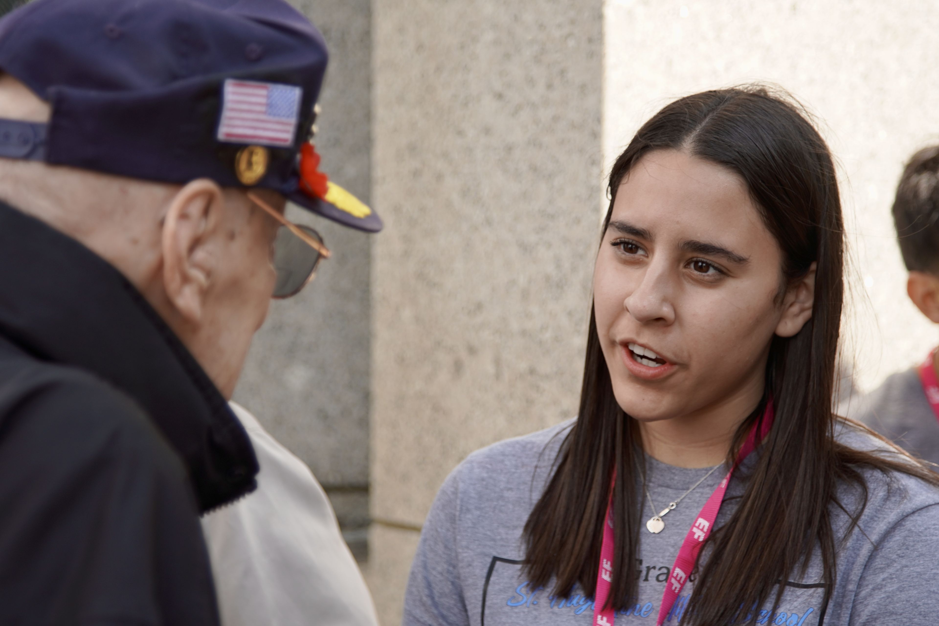 A young woman with long dark hair talks to an older man wearing a cap adorned with a small American flag.