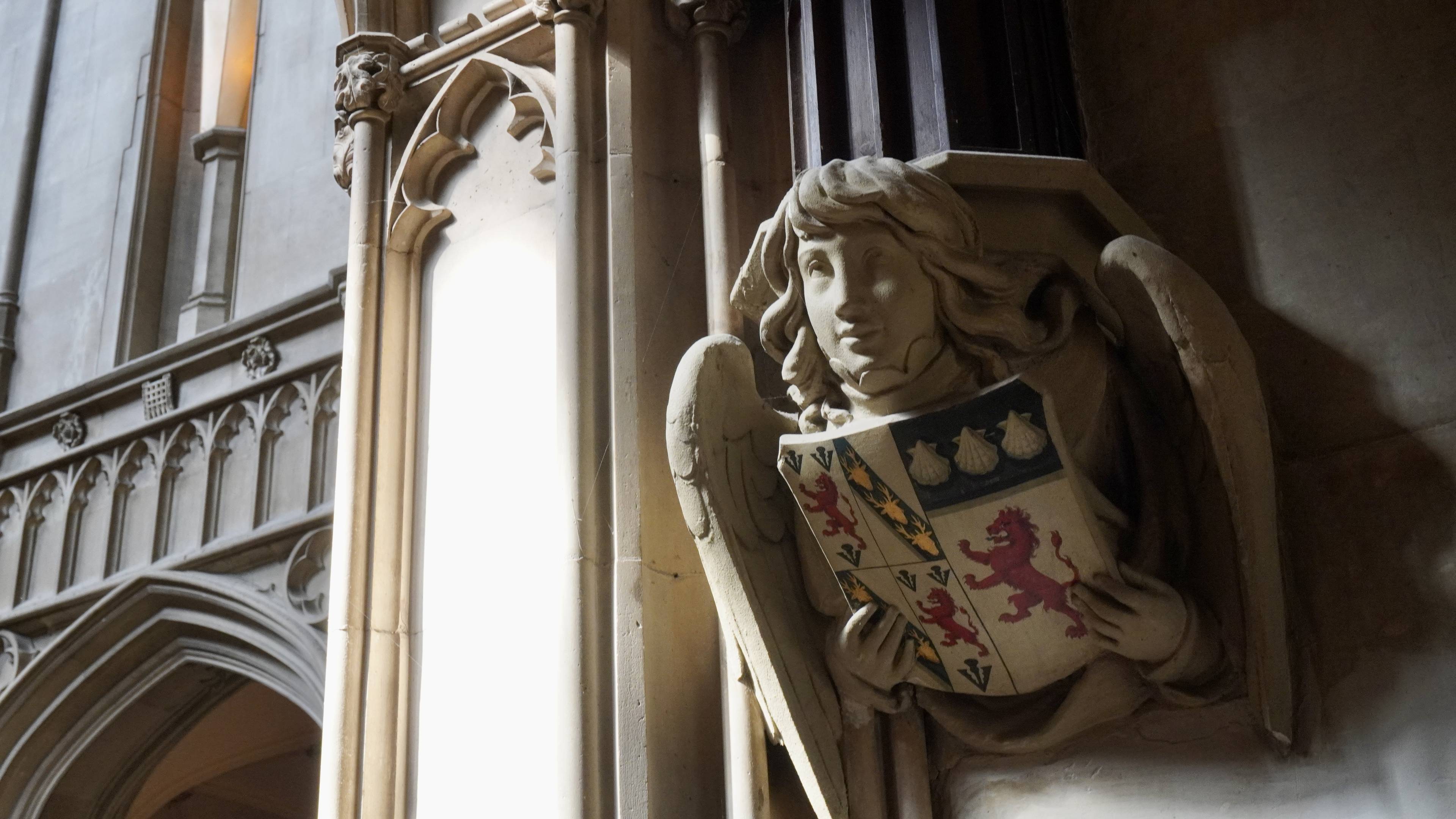 Stone angel sculpture holding a heraldic shield, set in a Gothic architectural interior with ornate arches and soft lighting.